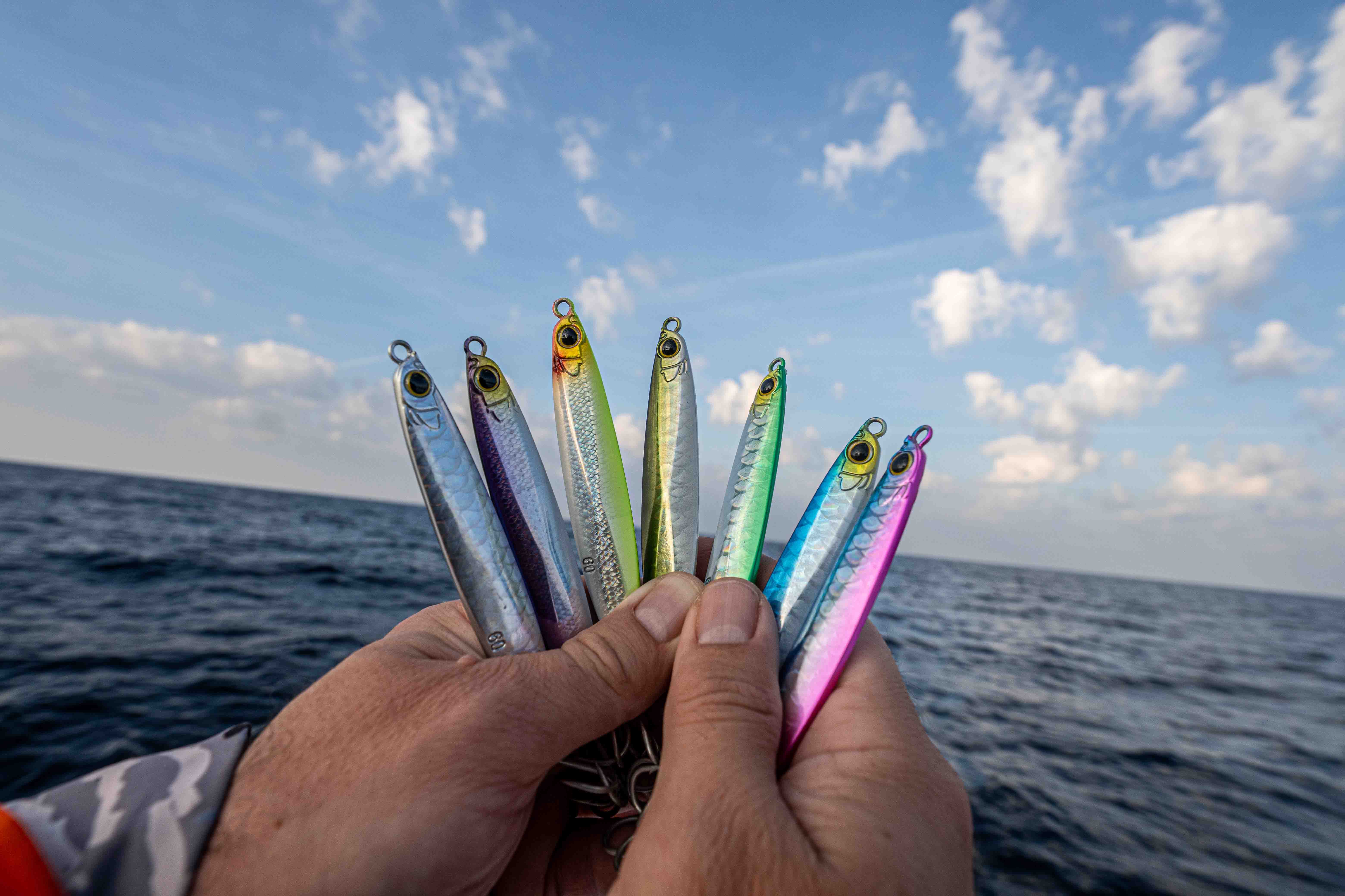 Hands holding seven metal jig fishing lures in silver-colored, green, blue, purple, and pink finishes above open ocean water under a blue sky with scattered clouds.