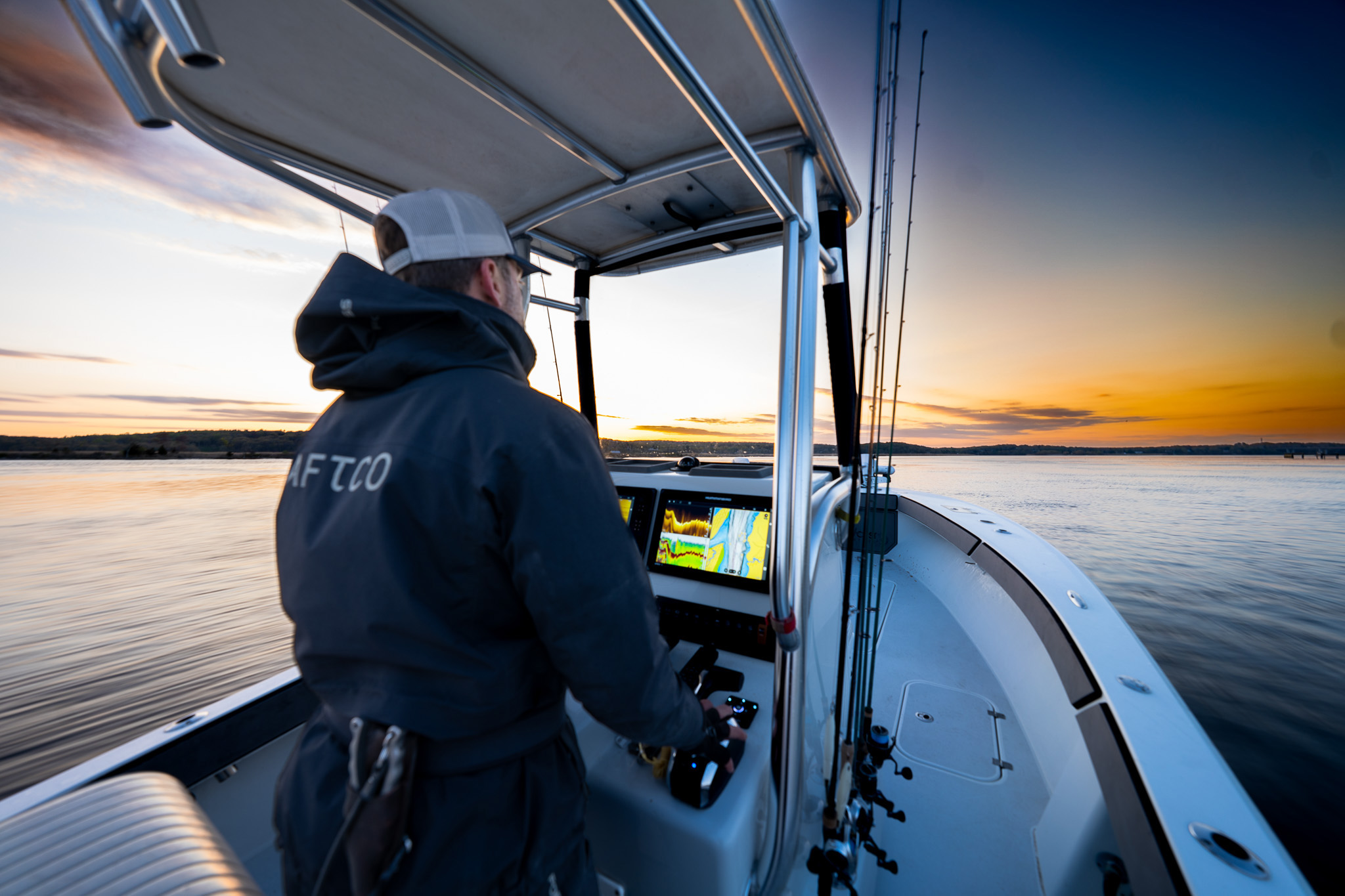 Person operating a center console fishing boat at sunrise with multiple fishing rods mounted near the helm, navigation screens visible, and calm water with an orange and blue sky in the background.