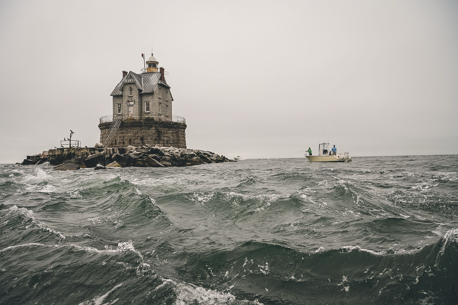 A small fishing boat with two people rides choppy ocean waves near a stone lighthouse on a rocky outcrop beneath an overcast sky.