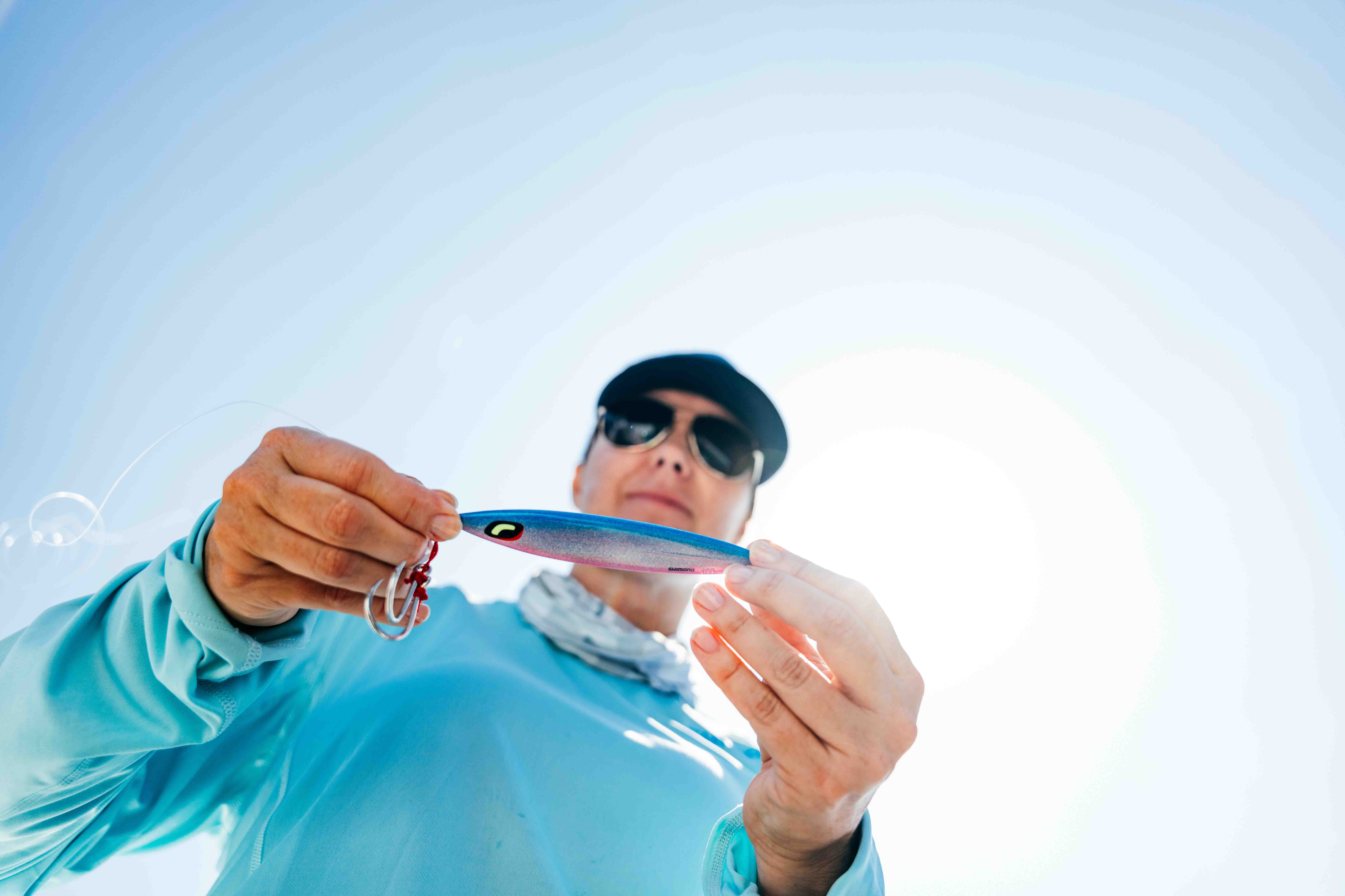 Person wearing sunglasses, a dark cap, and a light blue shirt holds a blue and pink metal jig lure with assist hooks toward the camera against a bright sky.
