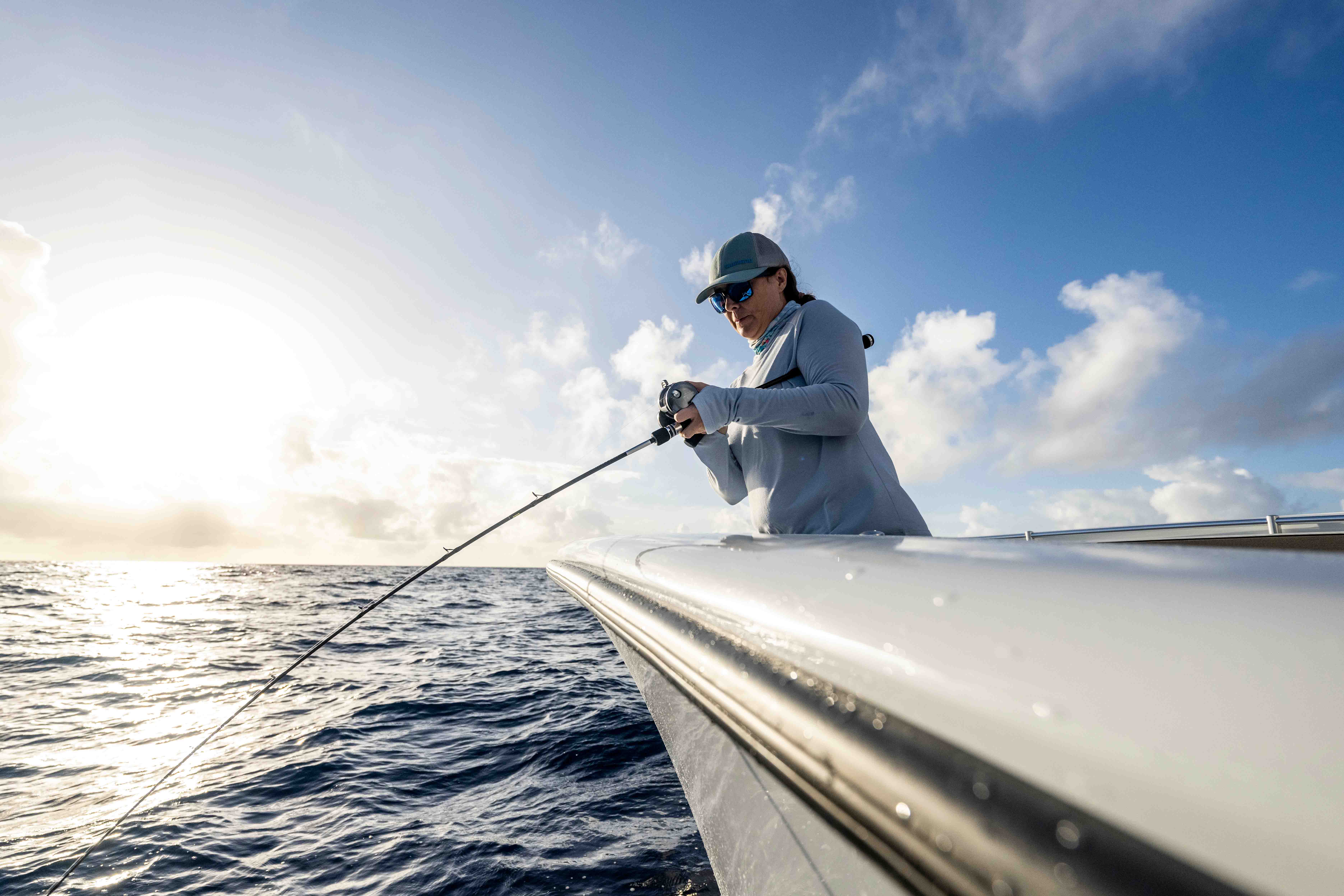 Woman standing on a boat above open ocean, holding a fishing rod and reel while looking toward the water, wearing a gray cap, blue mirrored sunglasses, and a light gray long-sleeve shirt, with clouds and low sun in the background.