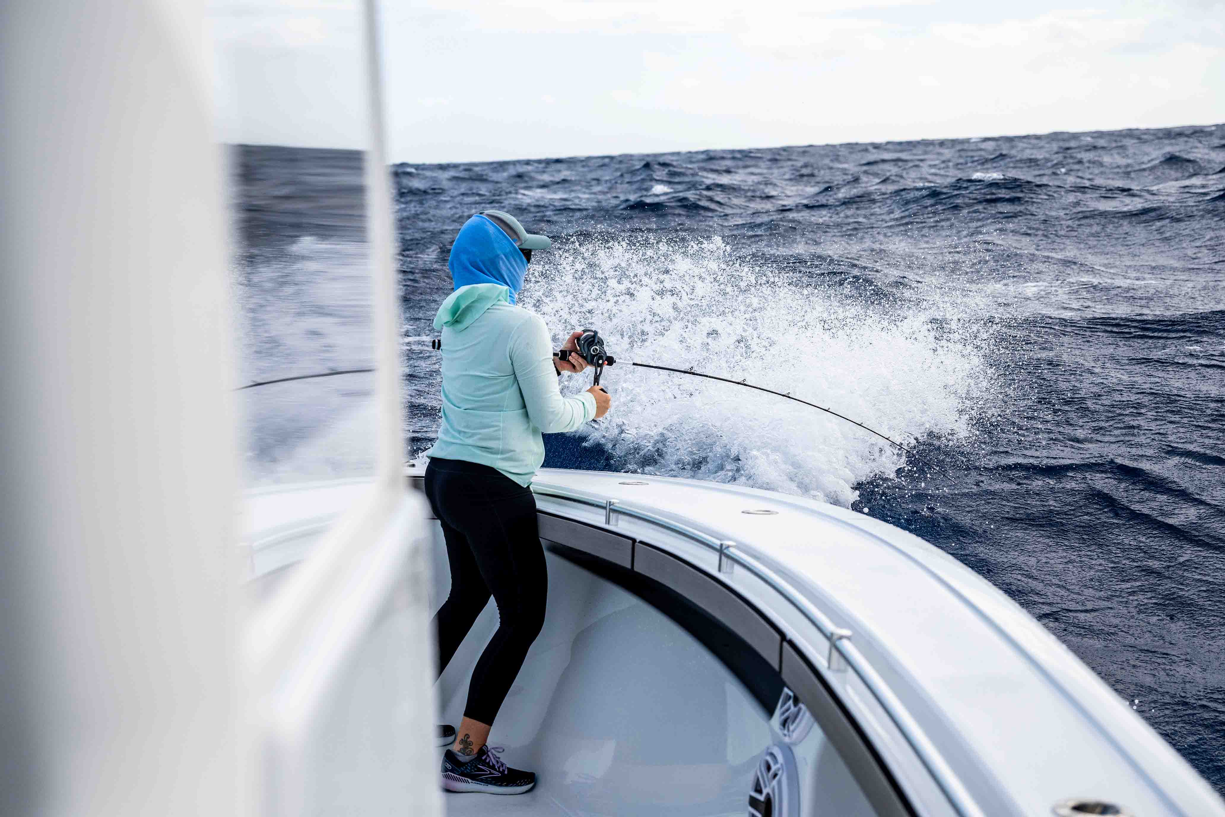 Woman fishing from the bow of a boat in rough ocean conditions, holding a bent fishing rod and reel as waves crash alongside the vessel, wearing a light blue hooded shirt, cap, and black leggings.