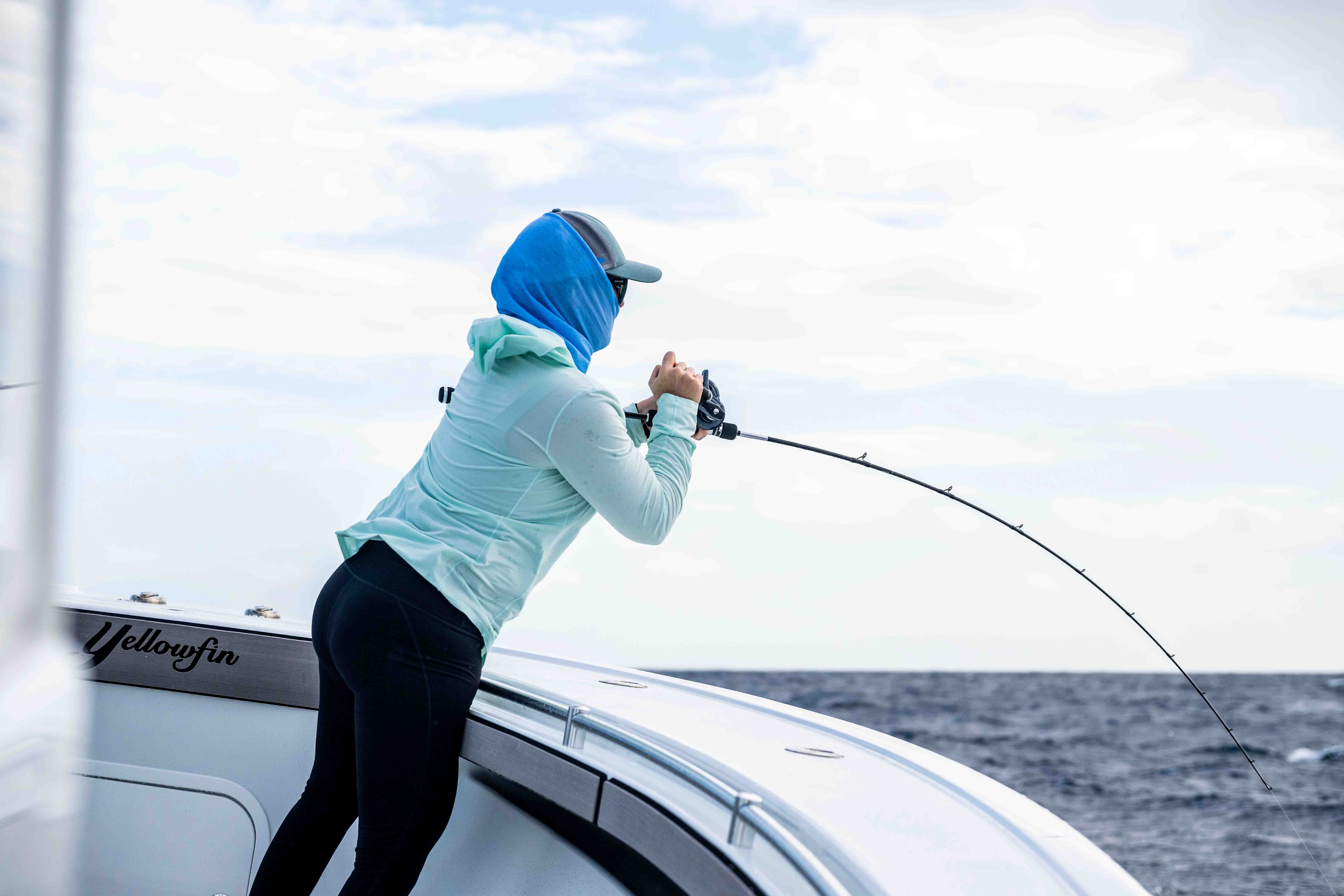 Woman standing on the bow of a boat offshore, fighting a fish with a bent fishing rod and reel, wearing a light blue hooded shirt, blue face covering, cap, and black leggings, with ocean and cloudy sky in the background.