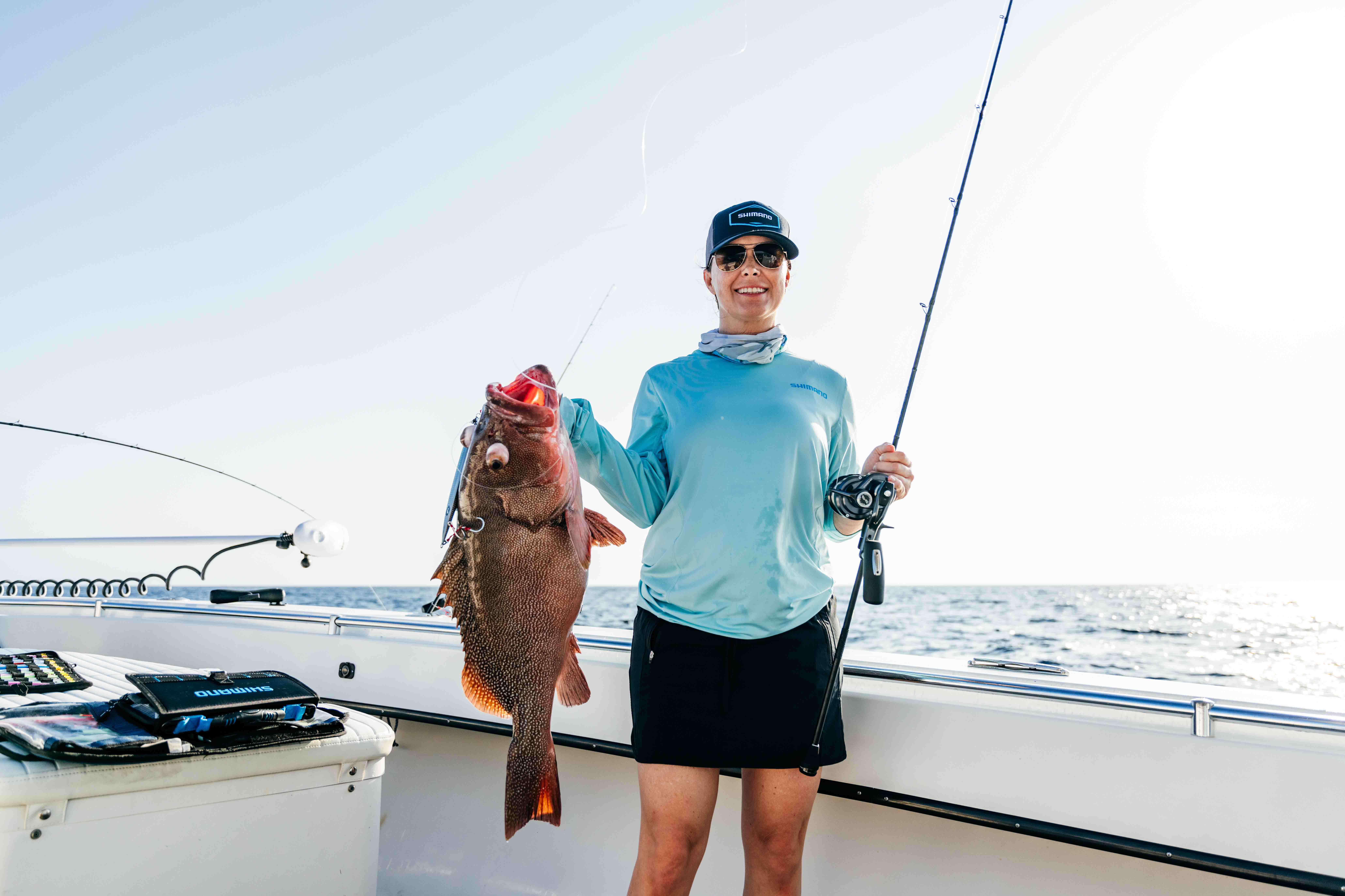 Woman standing on a boat holding a large reddish-brown fish in one hand and a fishing rod and reel in the other, wearing sunglasses, a dark cap with text "SHIMANO" in white text, and a light blue shirt, with the ocean in the background.