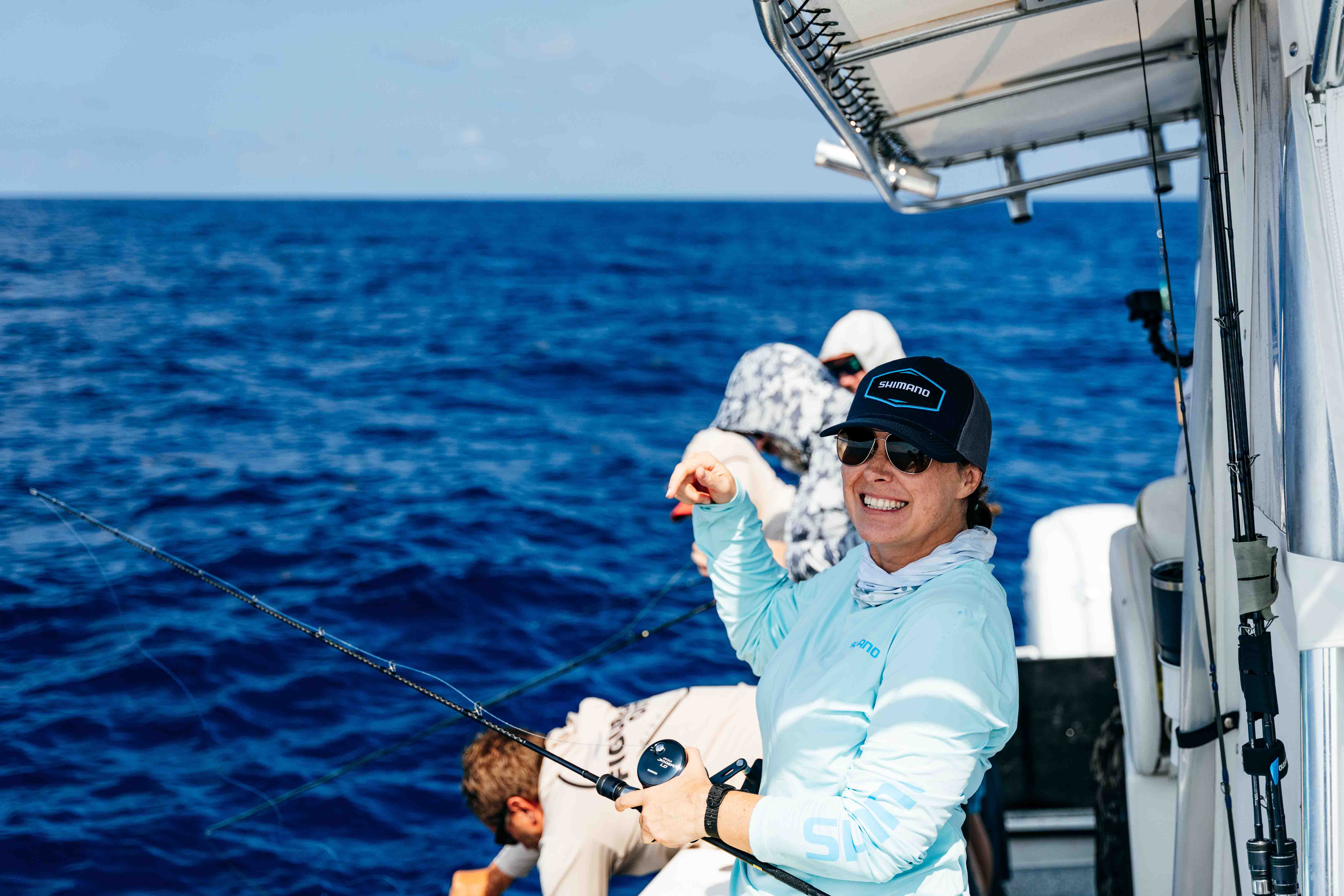 Woman wearing sunglasses, a dark cap with text "SHIMANO" in white text, and a light blue shirt holds a fishing rod on a boat, smiling toward the camera, with other anglers and open ocean in the background.
