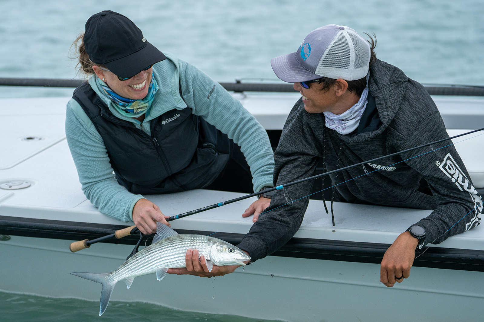 Two people are leaning over the side of a boat, one holding a silver fish and the other holding a fishing pole
