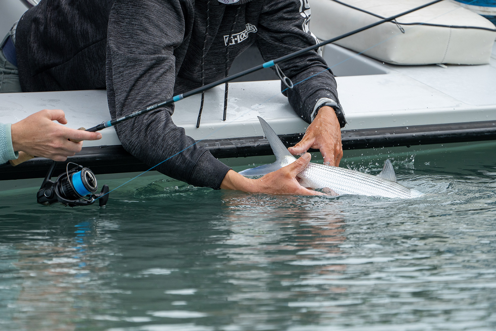 A person is leaning over the side of a boat while holding a silver fish. Another person is holding a blue and black fishing pole.
