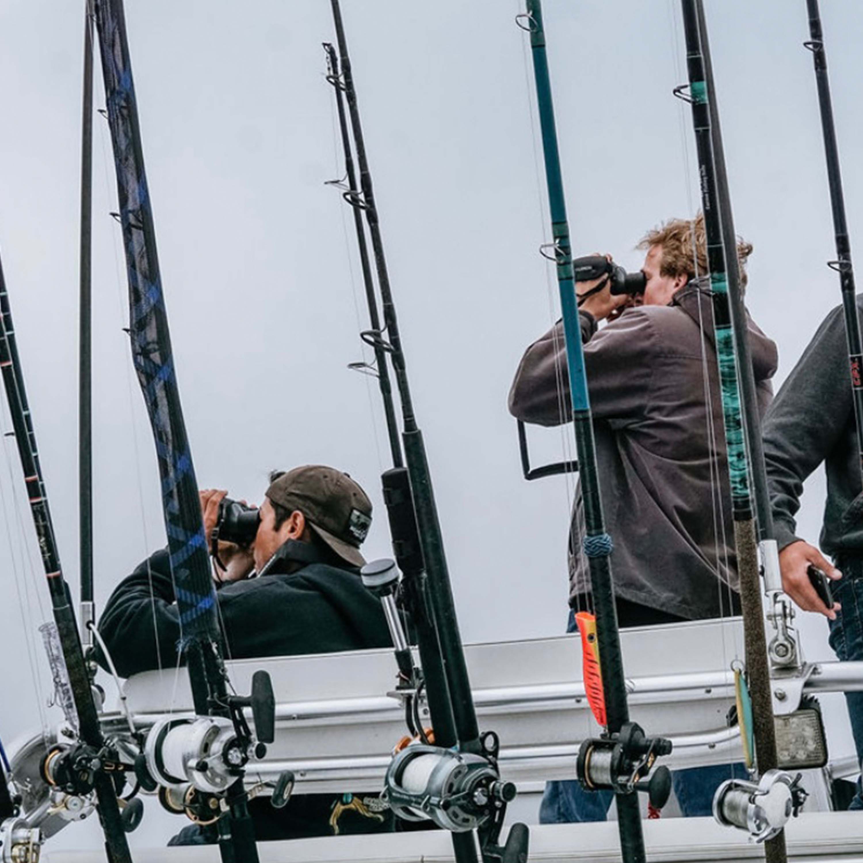 Two men stand watch on a boat tower, both using black gyroscopic binoculars. There are several rods and reels in the foreground, partially obstructing the men.