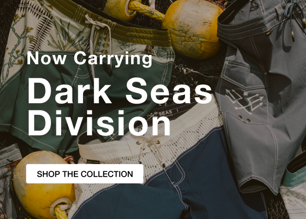 Promotional banner featuring Dark Seas Division board shorts and apparel arranged among fishing buoys and nets. Overlaid text reads, 'Now Carrying Dark Seas Division' with a button labeled 'Shop the Collection.