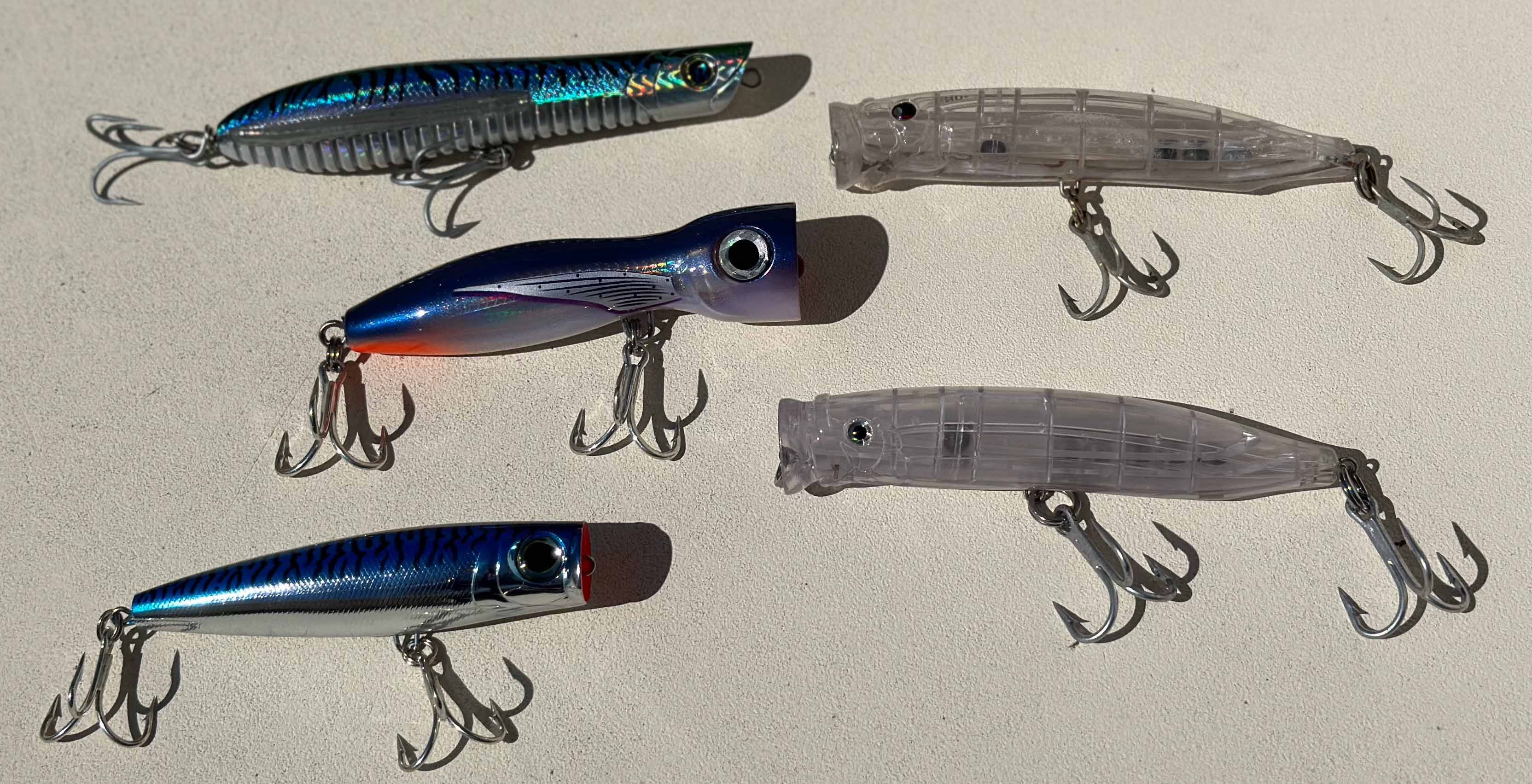 Five Treble Hooked Popper lures are on a textured surface. They are blue and white with various shapes.