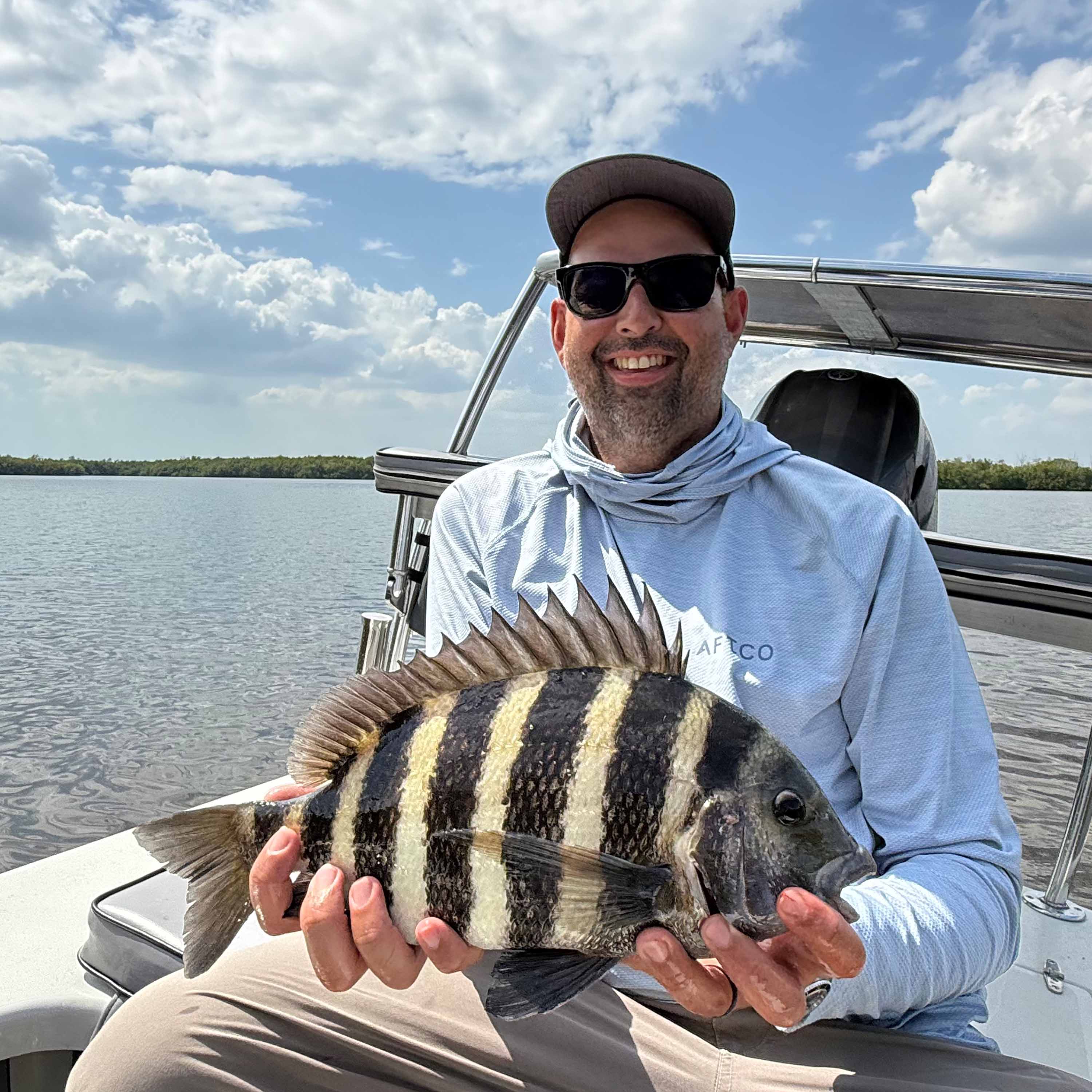 Andrew Alvarez sits on a boat holding a striped sheepshead fish, smiling on calm water under a partly cloudy sky.