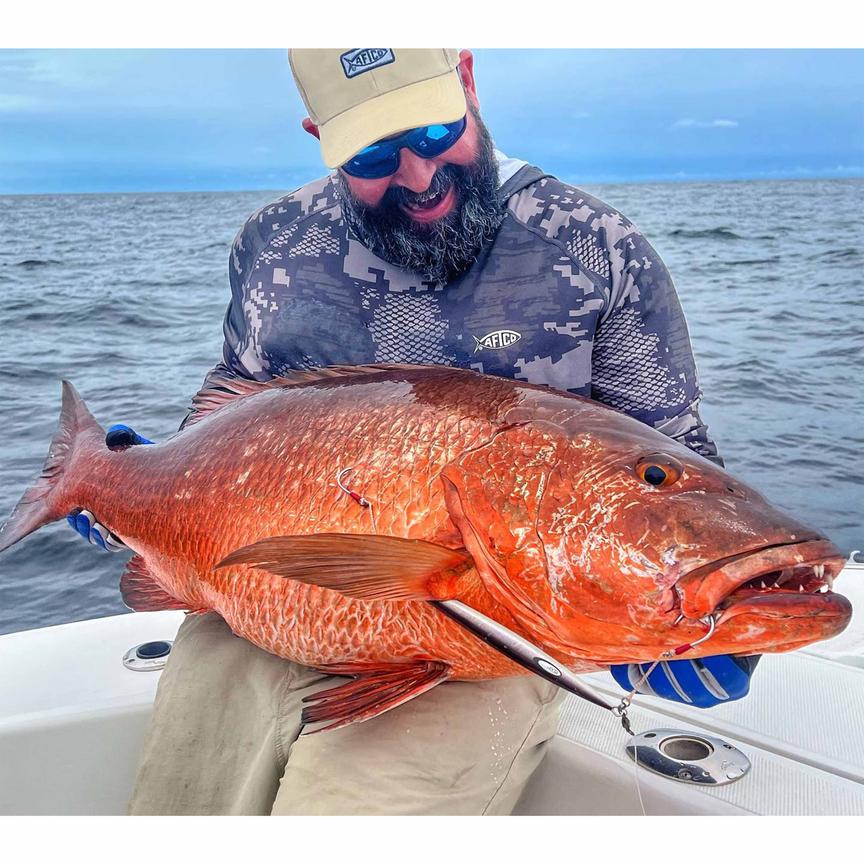 Benny Ortiz holds a large orange fish with a fishing lure in its mouth while seated on a boat on open water under an overcast sky.