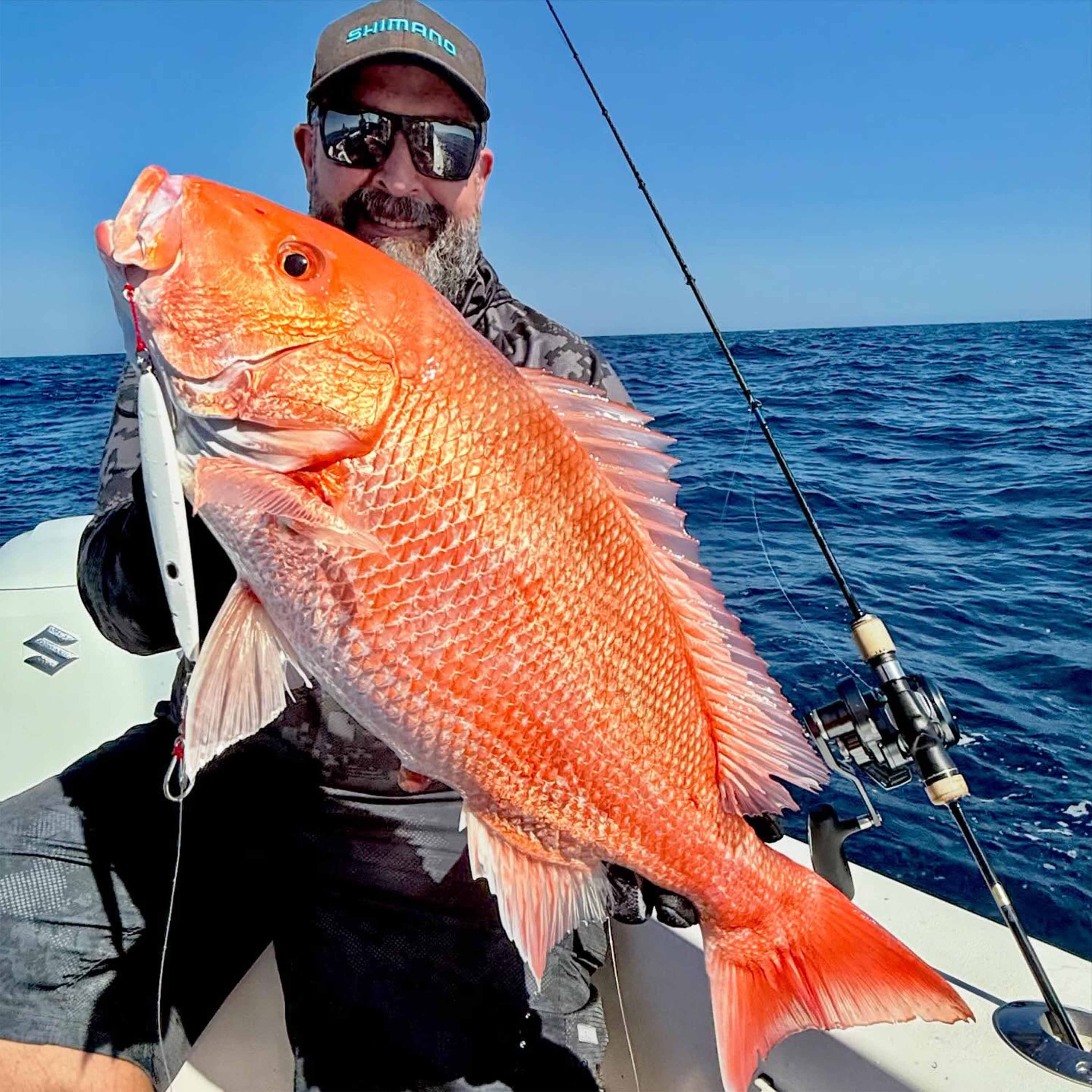 Benny Ortiz holds a large bright orange fish with a fishing lure in its mouth while aboard a boat on open water, with a fishing rod and clear blue sky in the background.