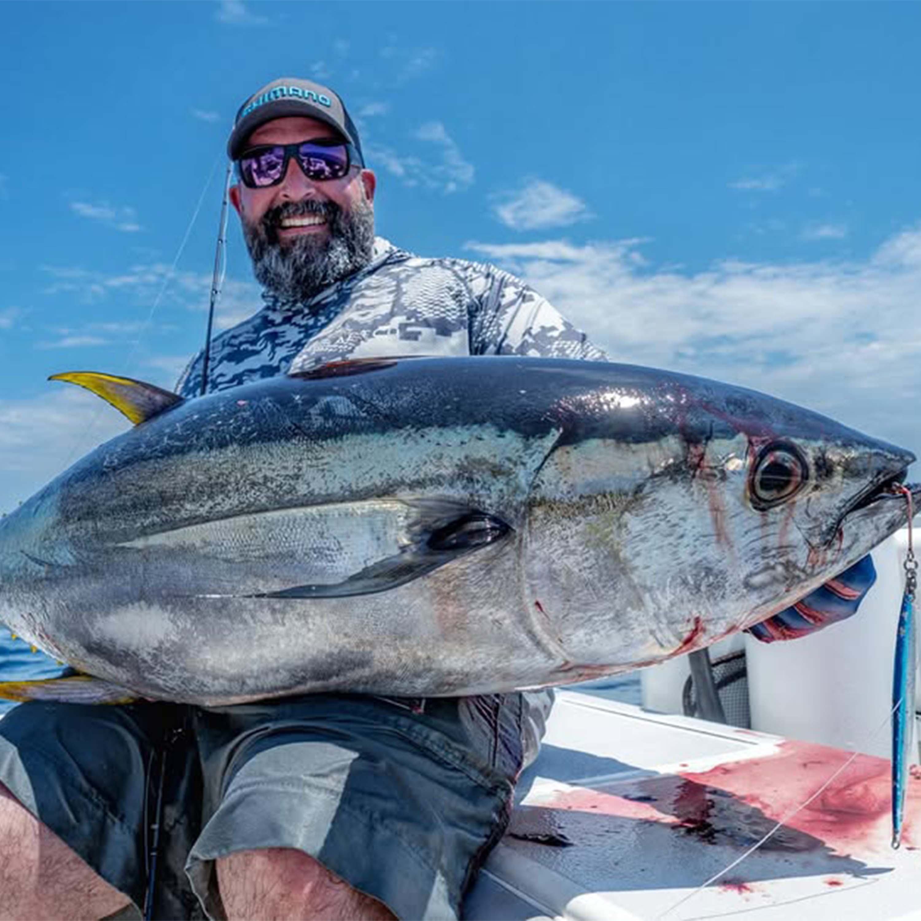 Benny Oriz smiles aboard a boat while holding a large tuna across his lap under a clear blue sky with a fishing lure visible near the fish's mouth.