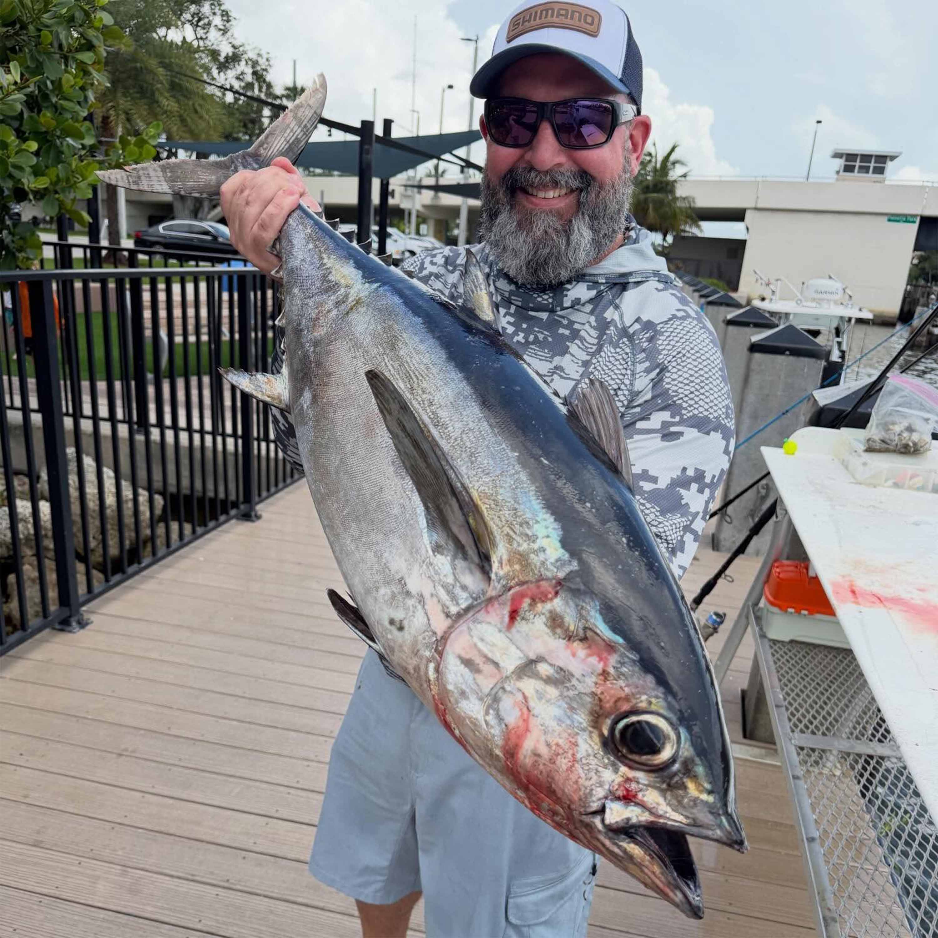 Benny Ortiz holds a large tuna while standing on a marina dock, with boats, railings, and waterfront buildings visible in the background.