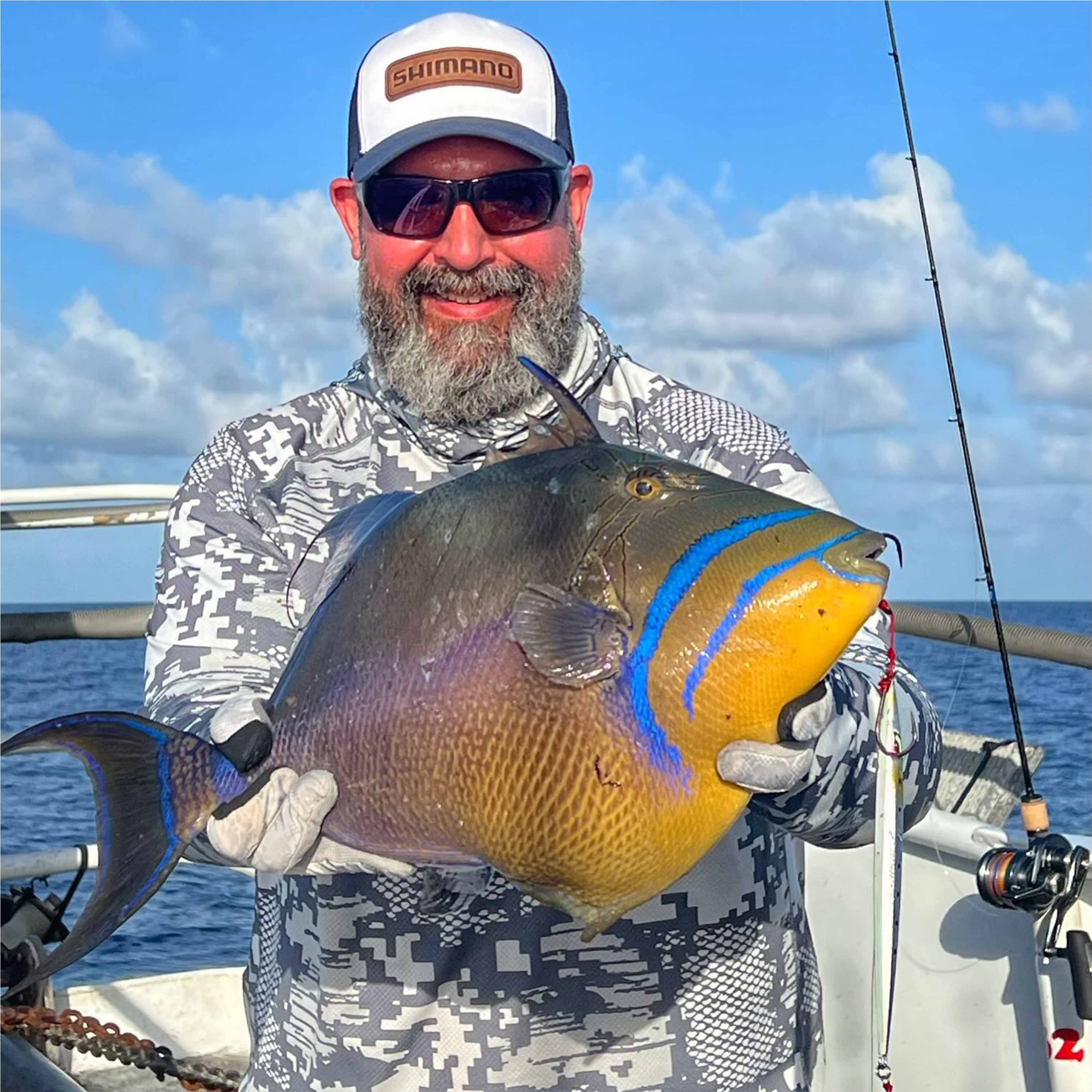 Benny Ortiz holds a colorful reef fish with blue markings and a yellow face on a boat at sea, with a fishing lure attached and a fishing rod visible against a cloudy blue sky.