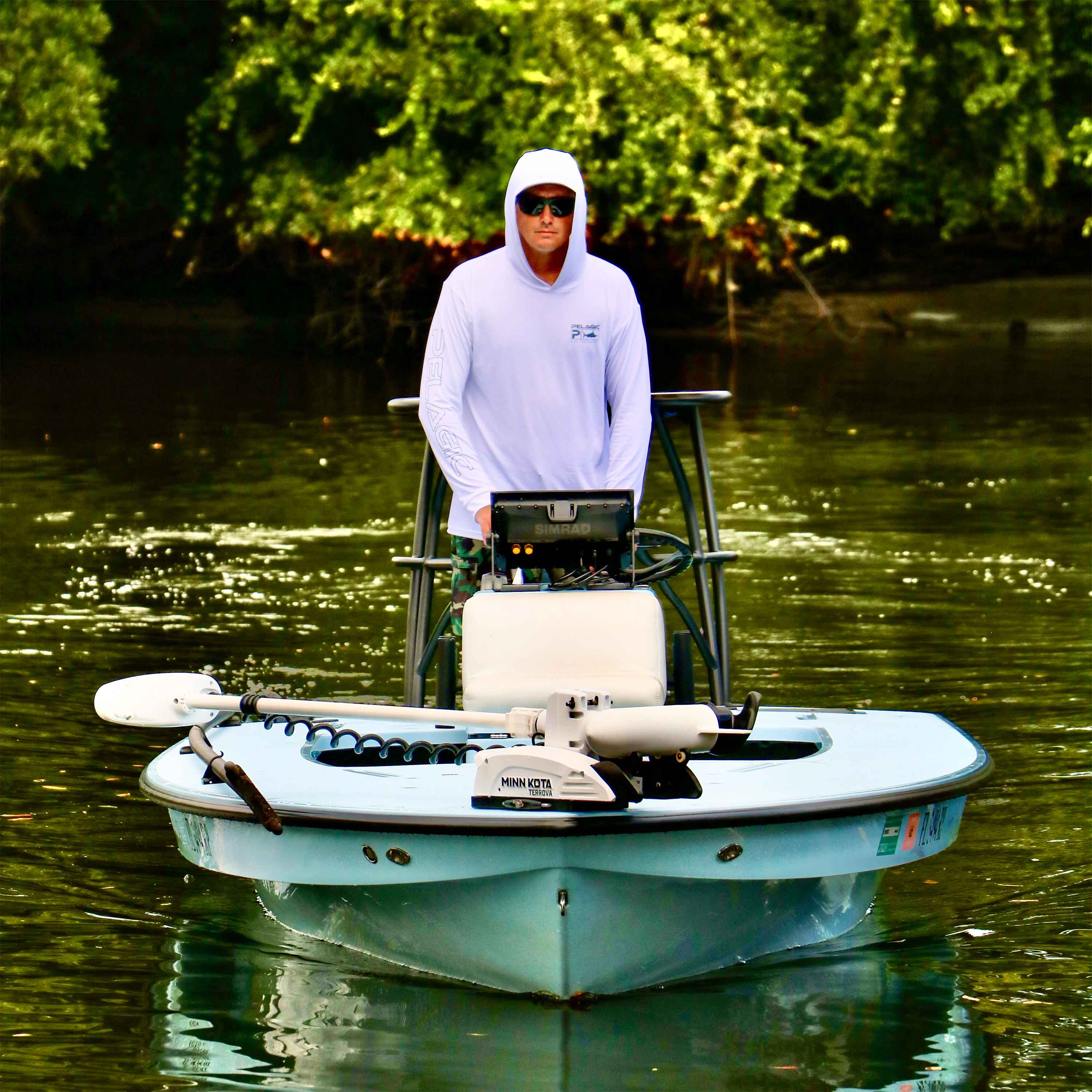Jarad Dingo Boshammer stands at the helm of a skiff on calm water surrounded by dense green trees.