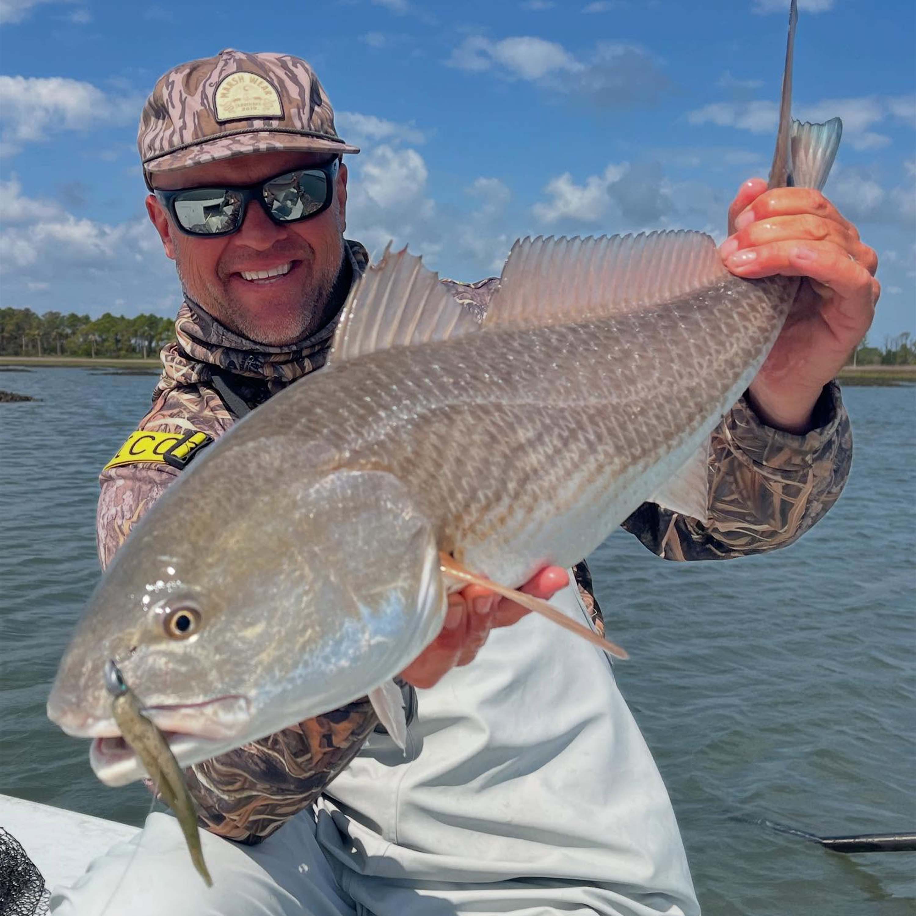 John Irwin holds a large fish on a boat, wearing camouflage fishing gear, sunglasses, and white bib overalls, with calm water and a blue sky in the background.