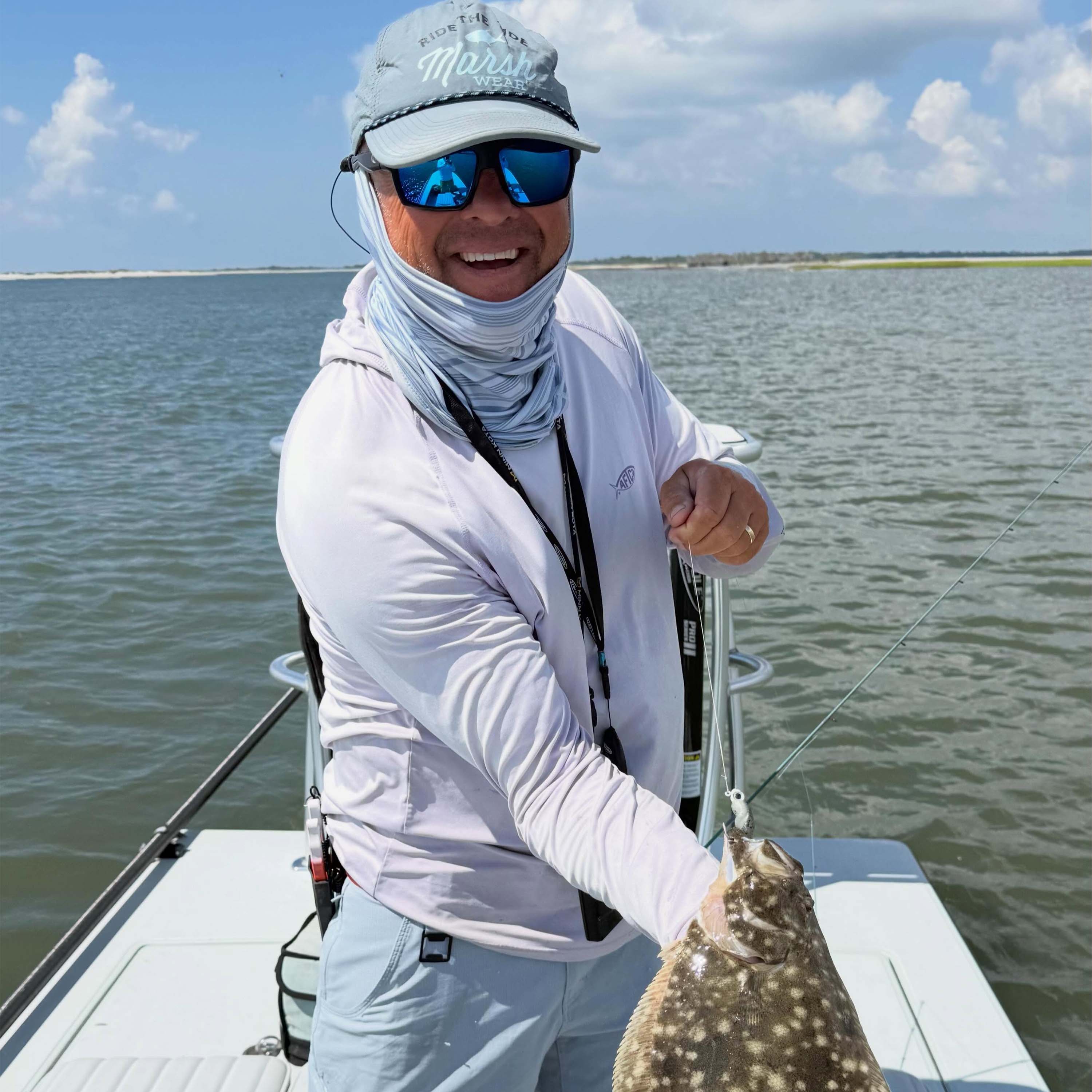 John Irwin stands on a boat holding a flatfish on a fishing line, wearing a white fishing shirt, face covering, sunglasses, and a light gray hat on calm water under a partly cloudy sky.