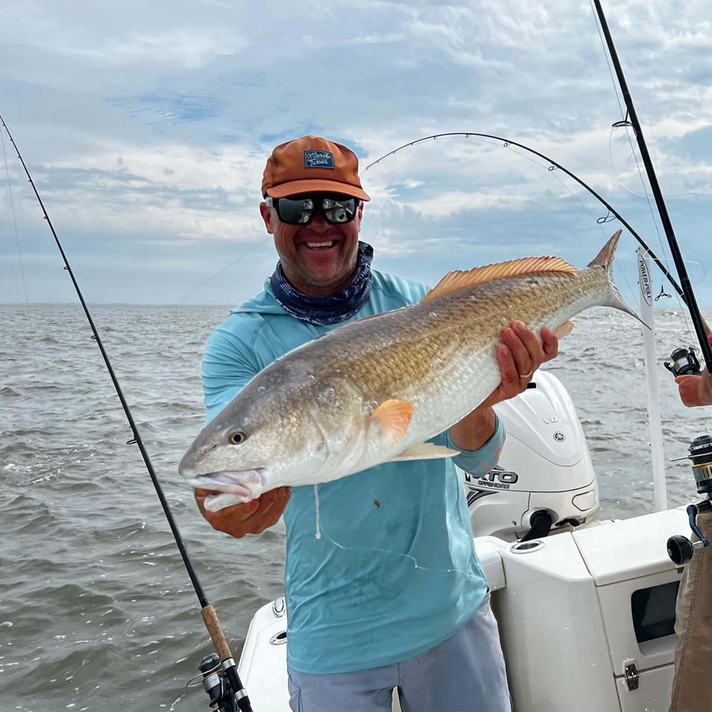 John Irwin holds a large fish on a boat, wearing a light blue fishing shirt, sunglasses, and an orange hat, with fishing rods, open water, and a cloudy sky in the background.