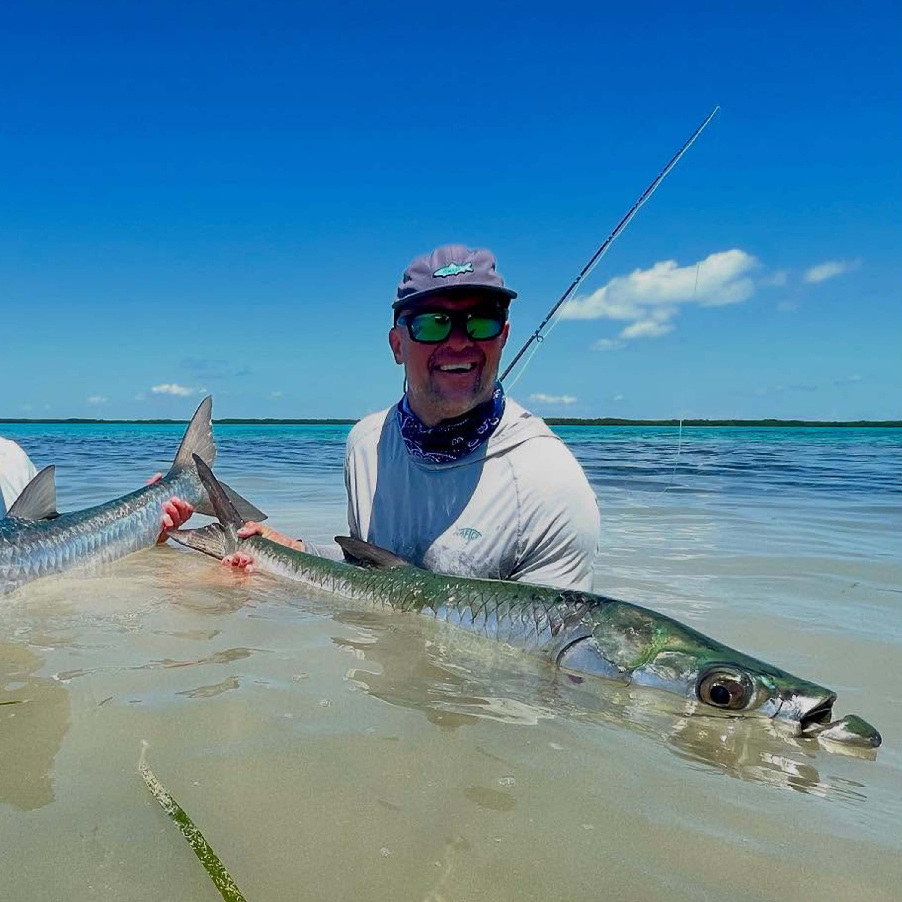 John Irwin kneels in shallow clear water while holding a long, slender fish, with another fish partially visible beside him under a bright blue sky.
