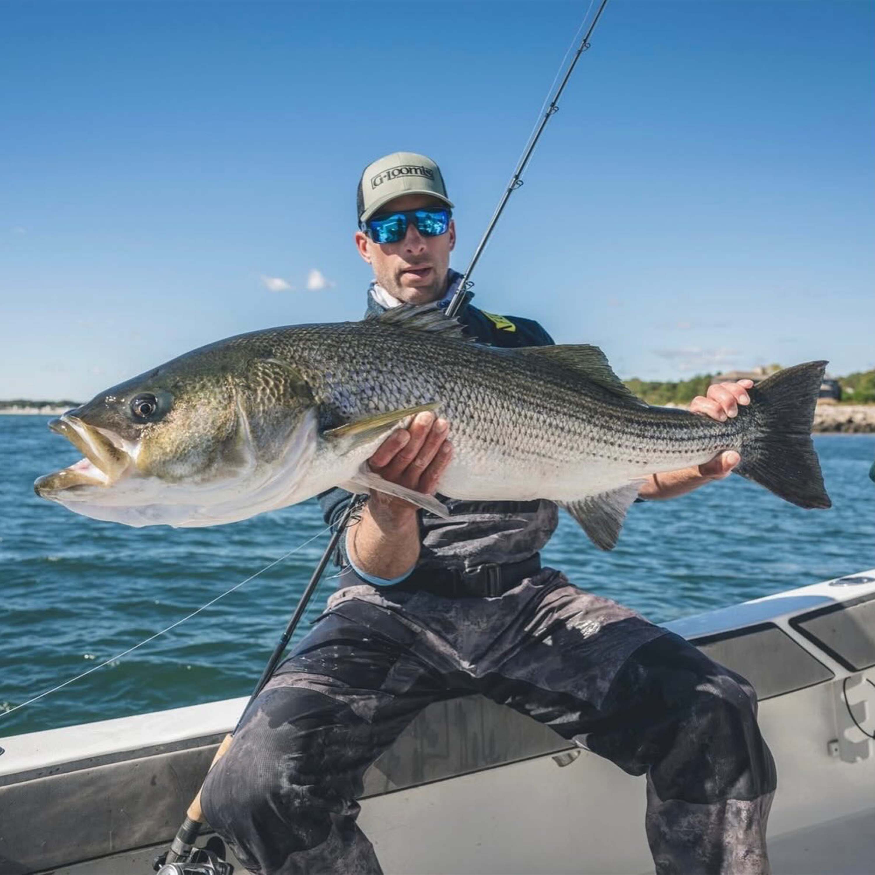 Captain Mike Roy holding a large striped bass aboard a fishing boat on Connecticut coastal waters under a clear blue sky.