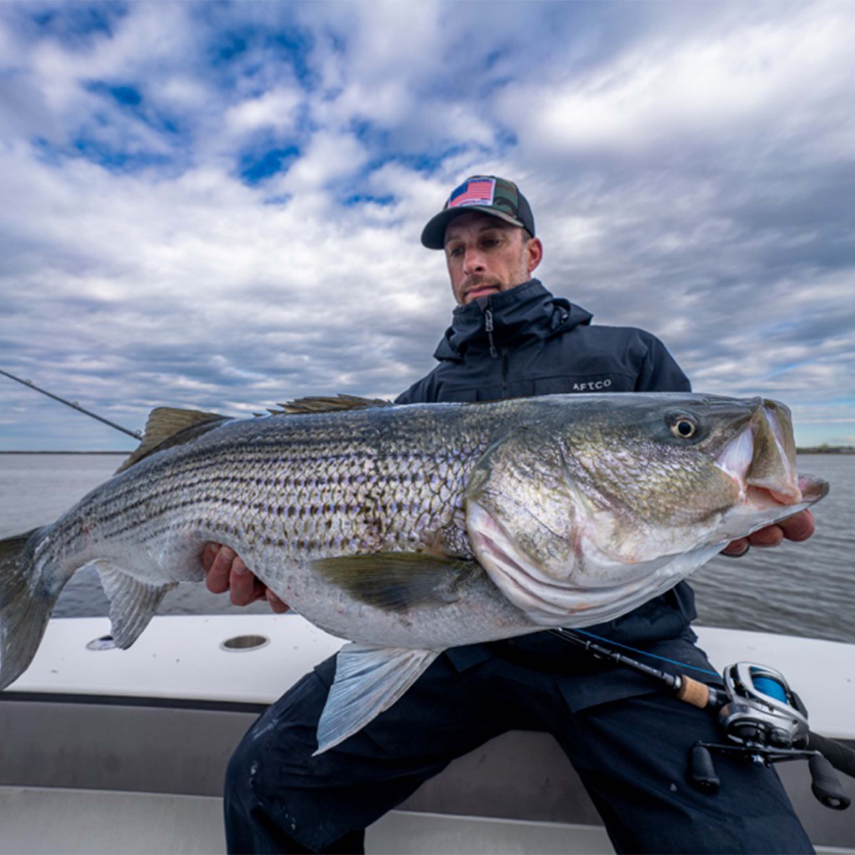 Captain Mike Roy holding a large striped bass aboard a fishing boat on Connecticut waters beneath a cloudy sky.
