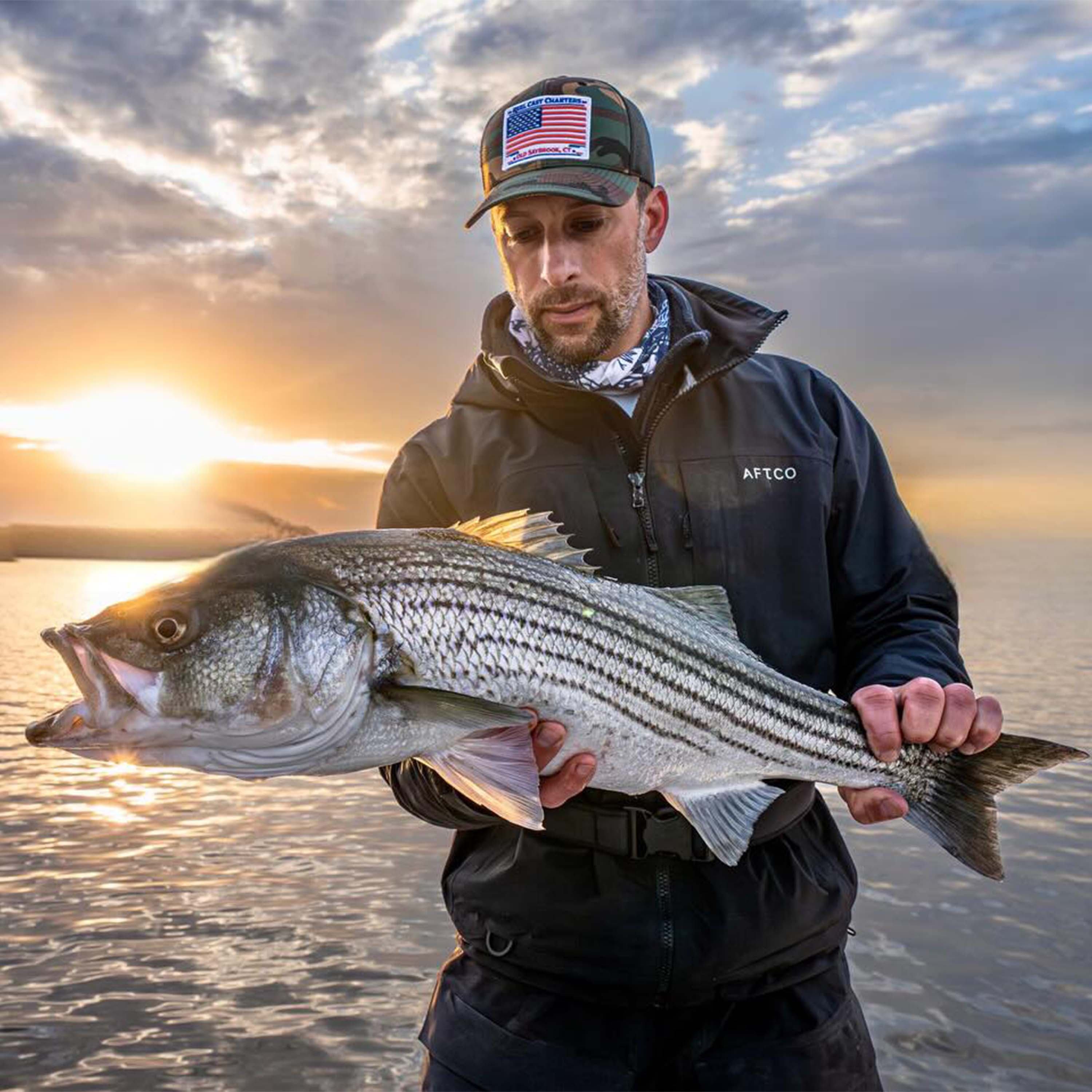 Mike Roy holds a striped bass over calm water at sunset, wearing a dark jacket and camouflage hat beneath a cloudy sky.