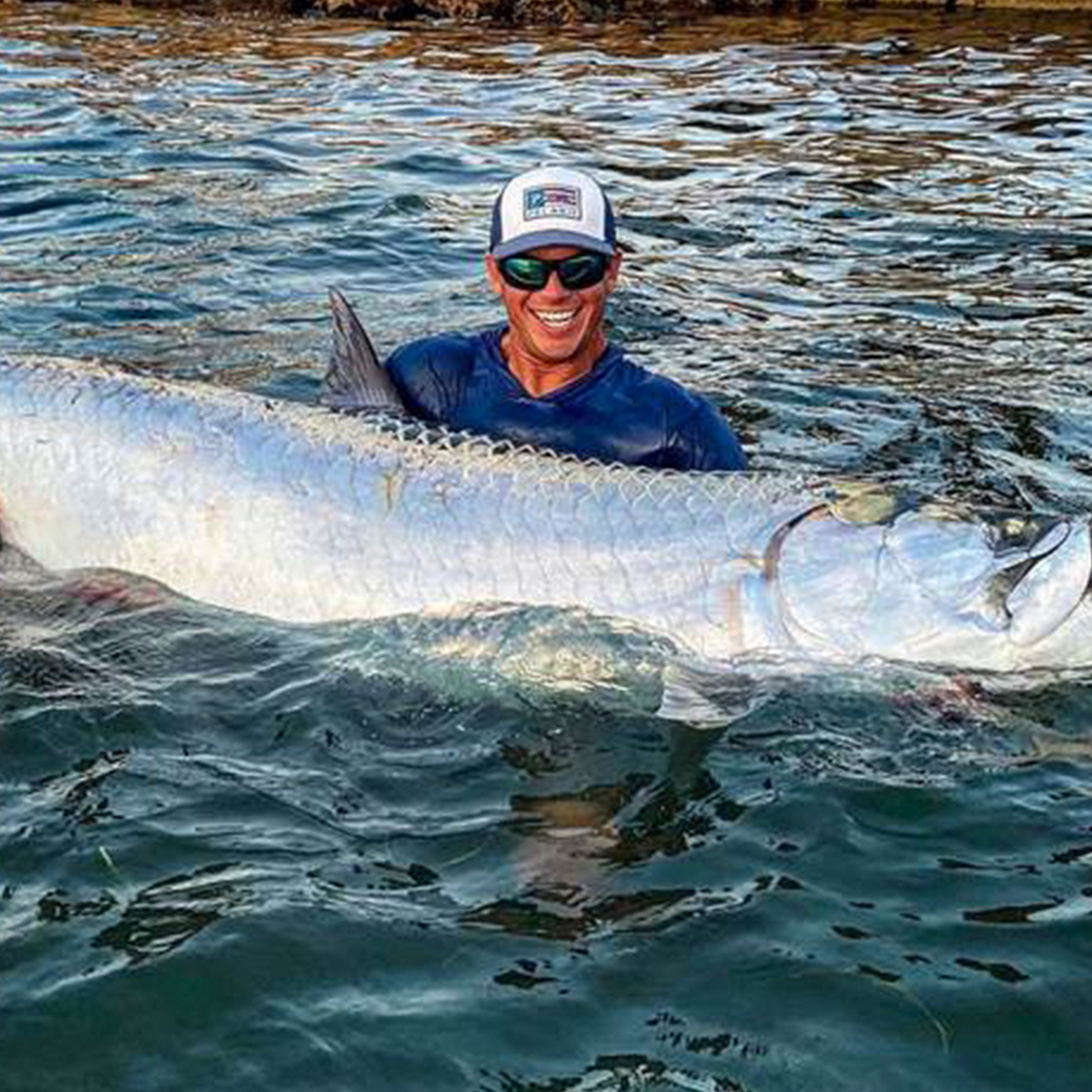 Tyler Kapela stands in the water holding a large silver fish, wearing a blue shirt, sunglasses, and a white hat, with rippling water surrounding them.
