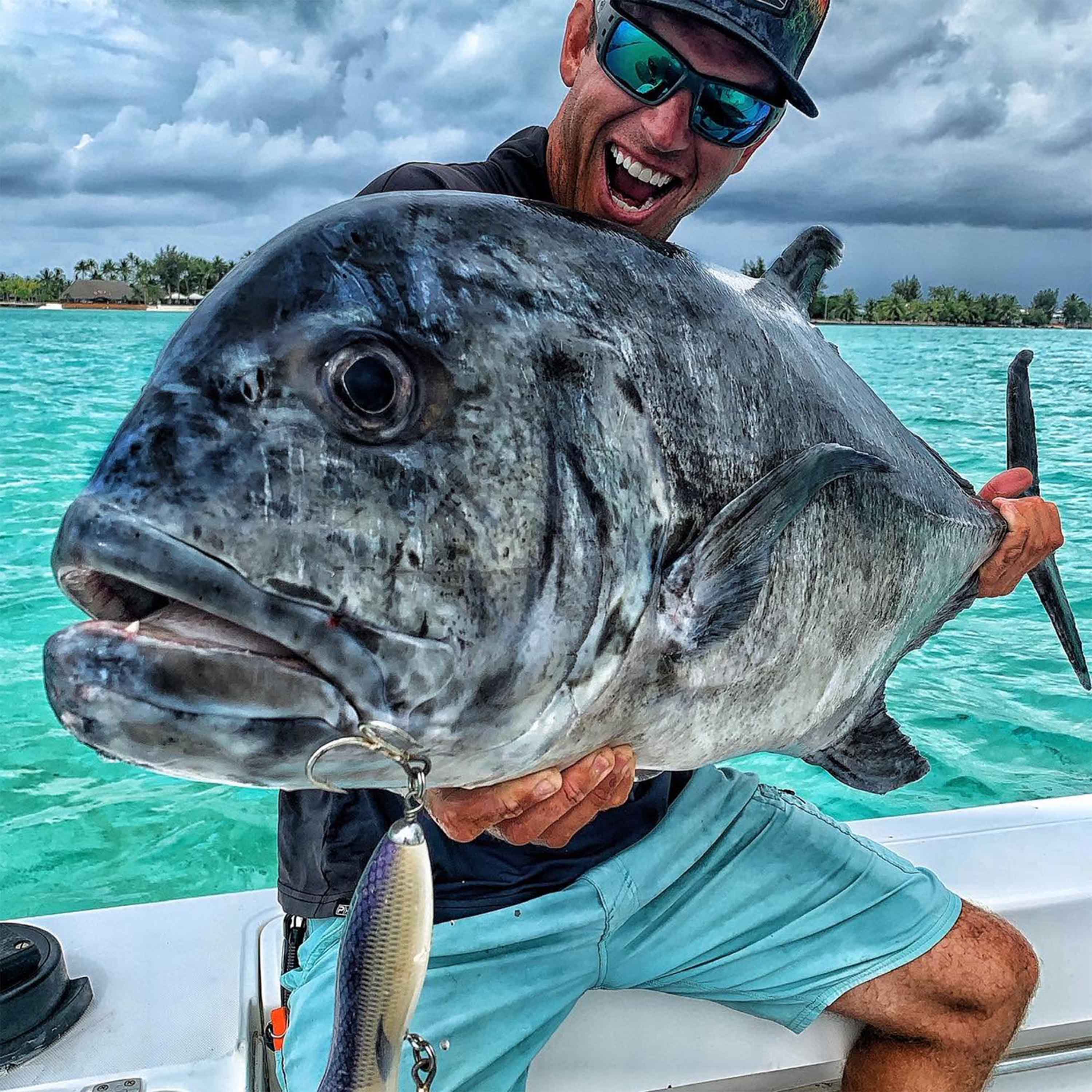 Tyler Kapela holds a large fish with a metal lure attached while sitting on a boat over clear turquoise water beneath a cloudy sky.