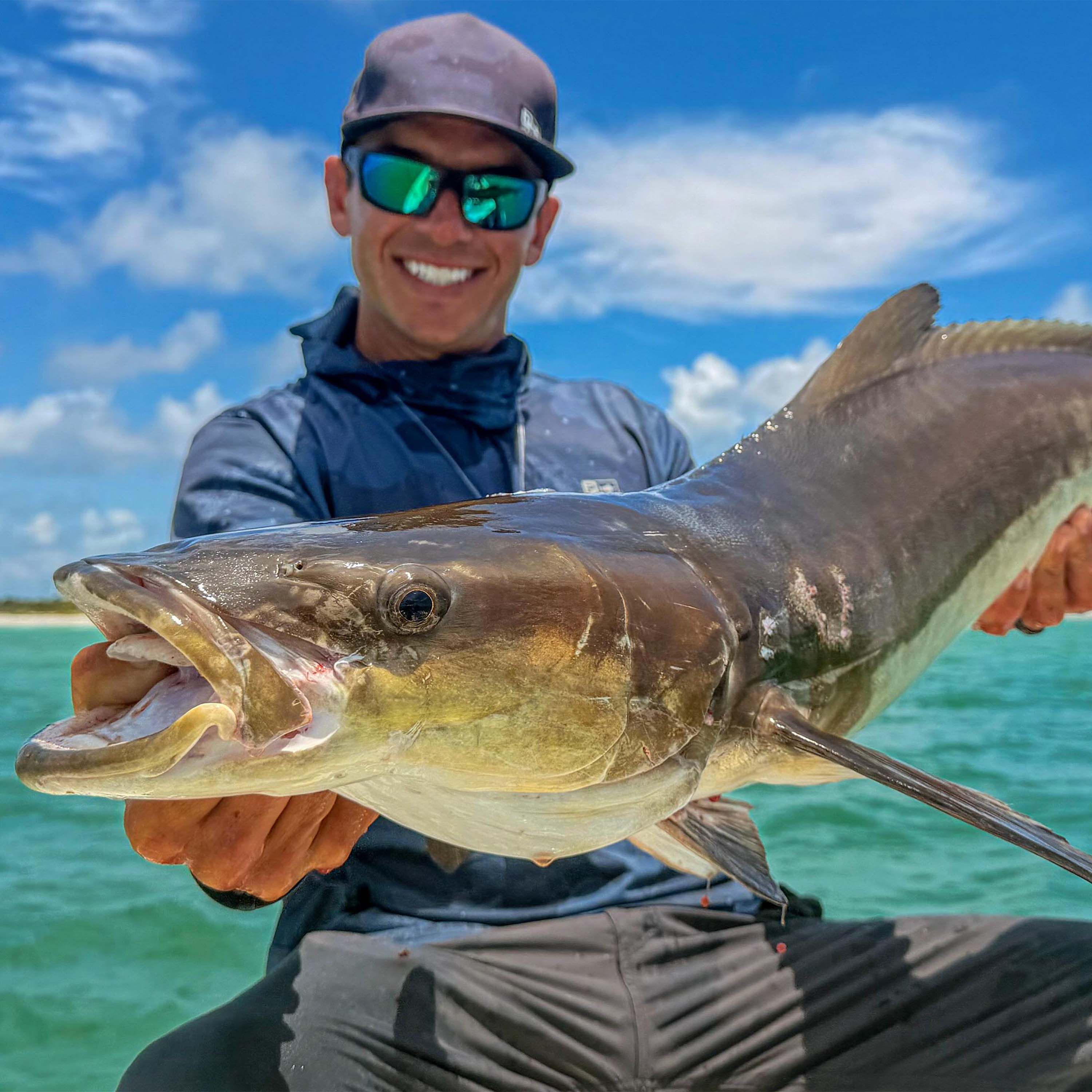 Tyler Kapela holds a large fish with a soft plastic lure in its mouth while on a boat over clear blue water beneath a bright, partly cloudy sky.