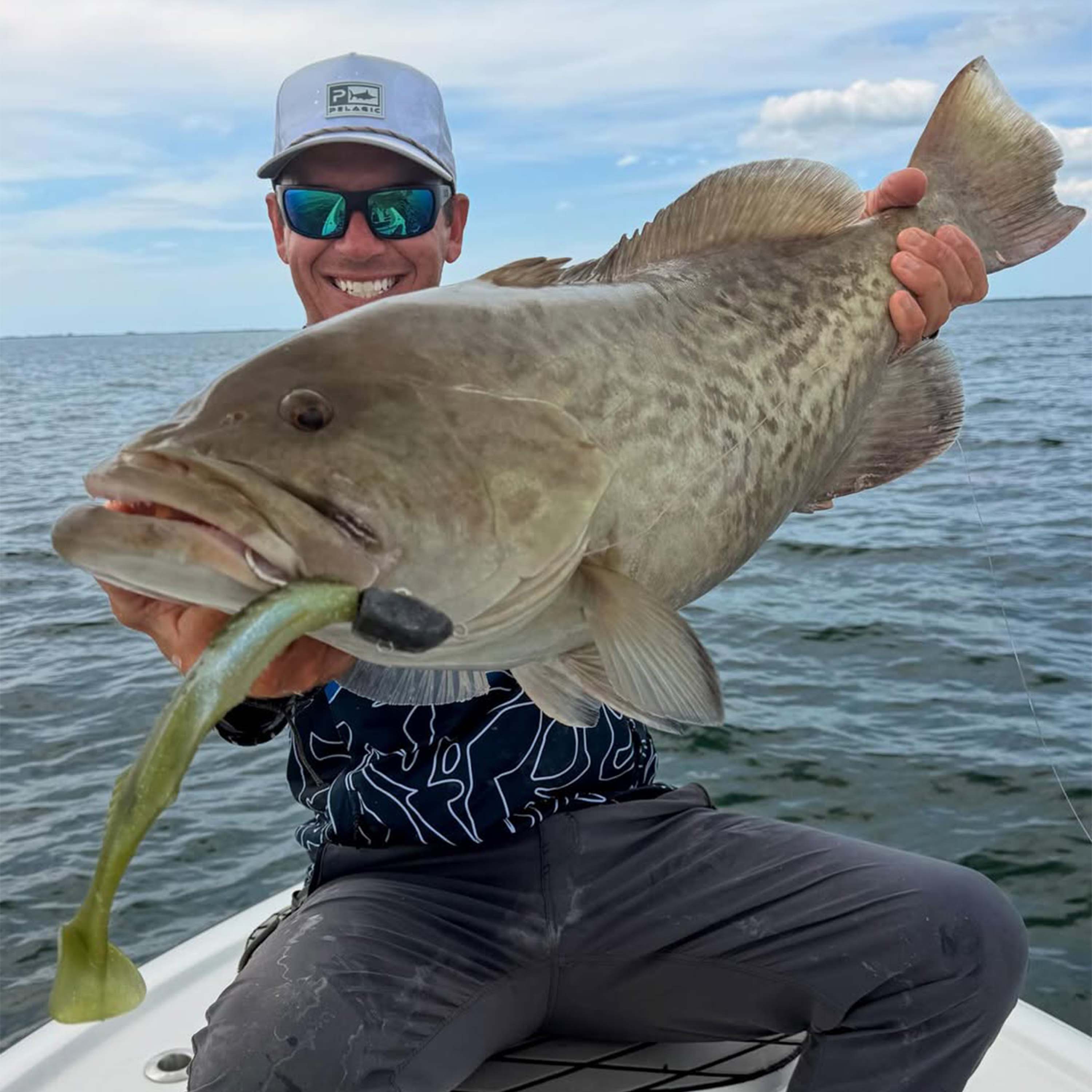 Tyler Kapela holds a large fish with a soft plastic lure in its mouth while sitting on a boat over open water beneath a partly cloudy sky.