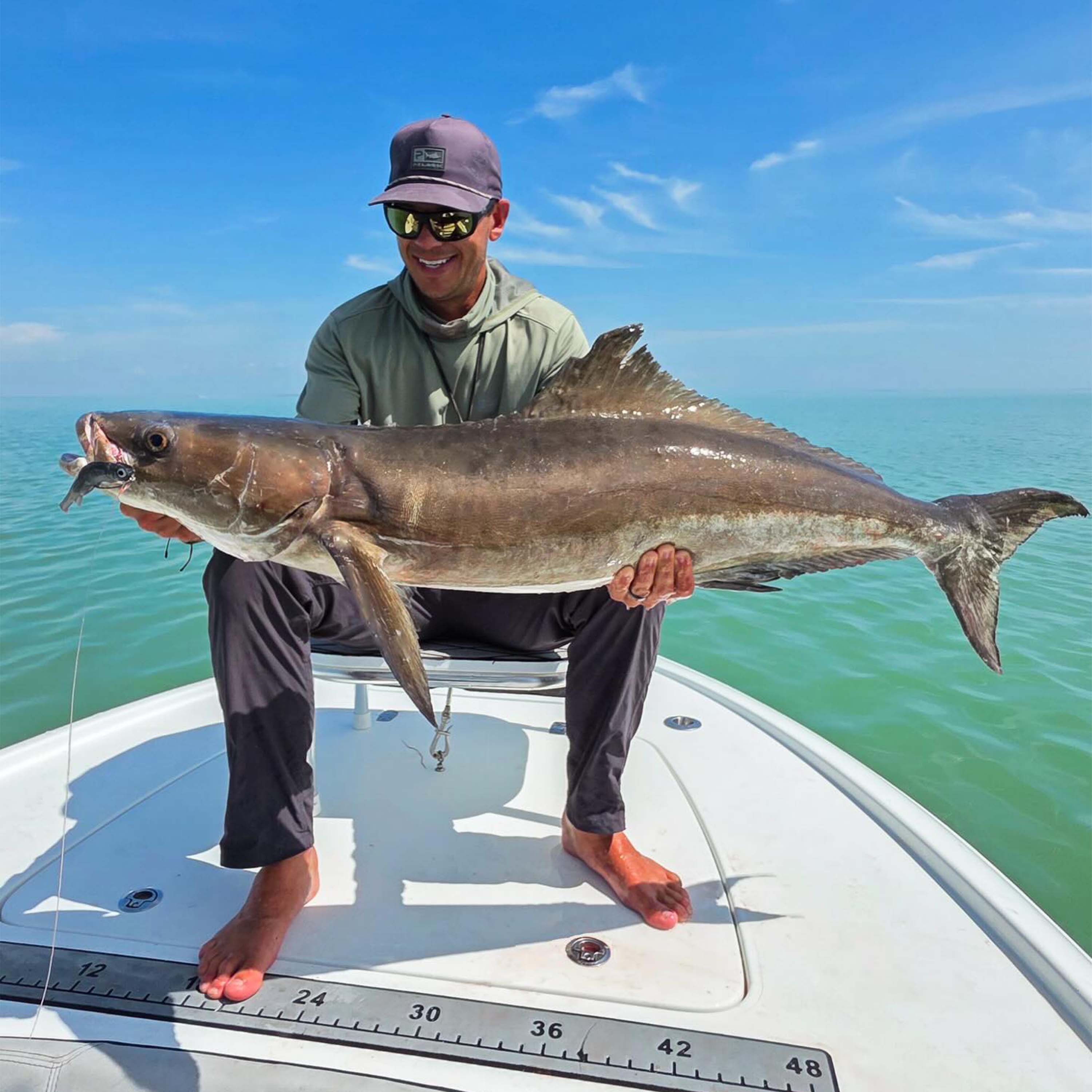 Tyler Kapela holds a large fish with a soft plastic lure in its mouth while sitting barefoot on the bow of a boat over calm turquoise water beneath a clear blue sky.