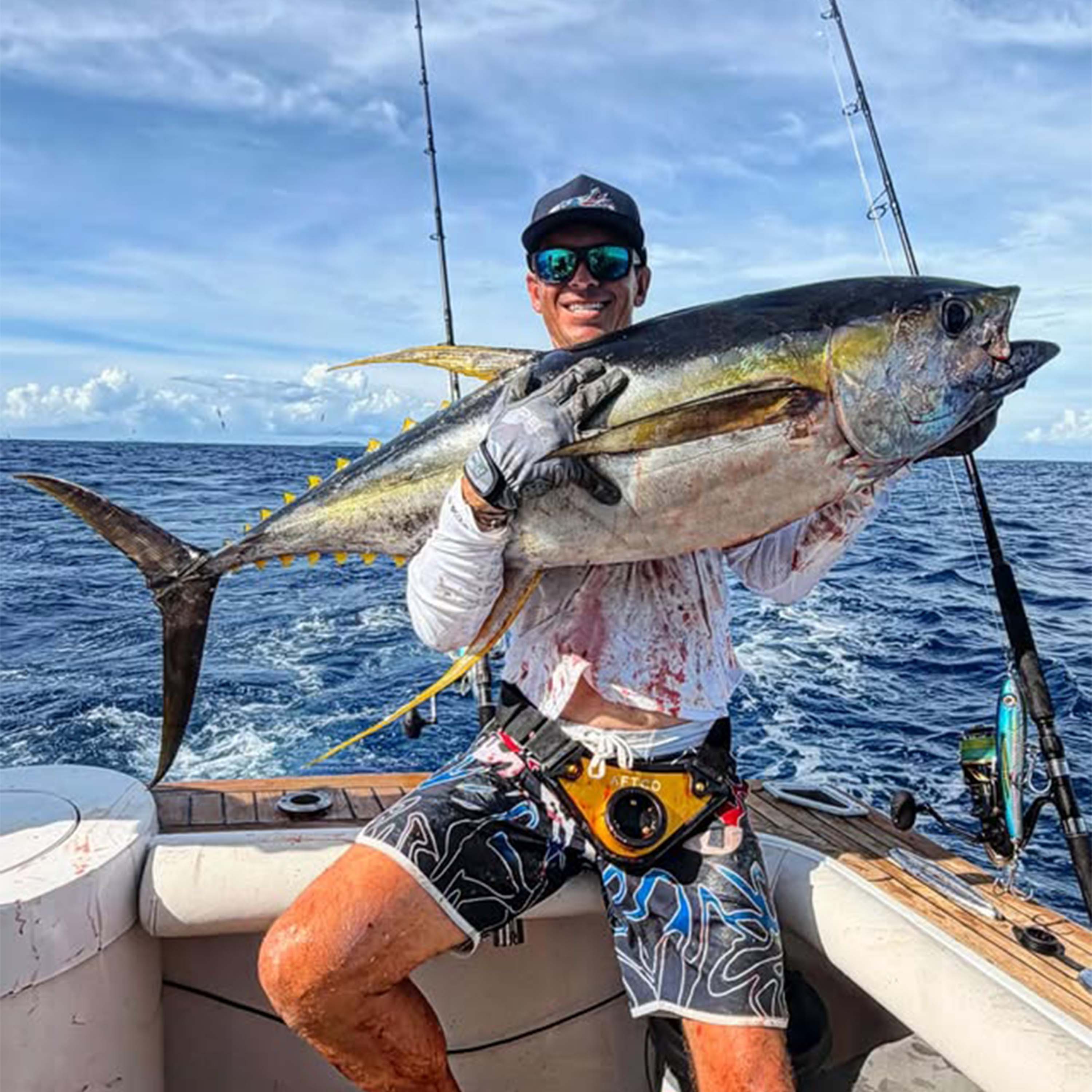 Tyler Kapela holds a large tuna on a sportfishing boat, wearing gloves, sunglasses, and fishing gear, with fishing rods and open ocean beneath a partly cloudy sky.
