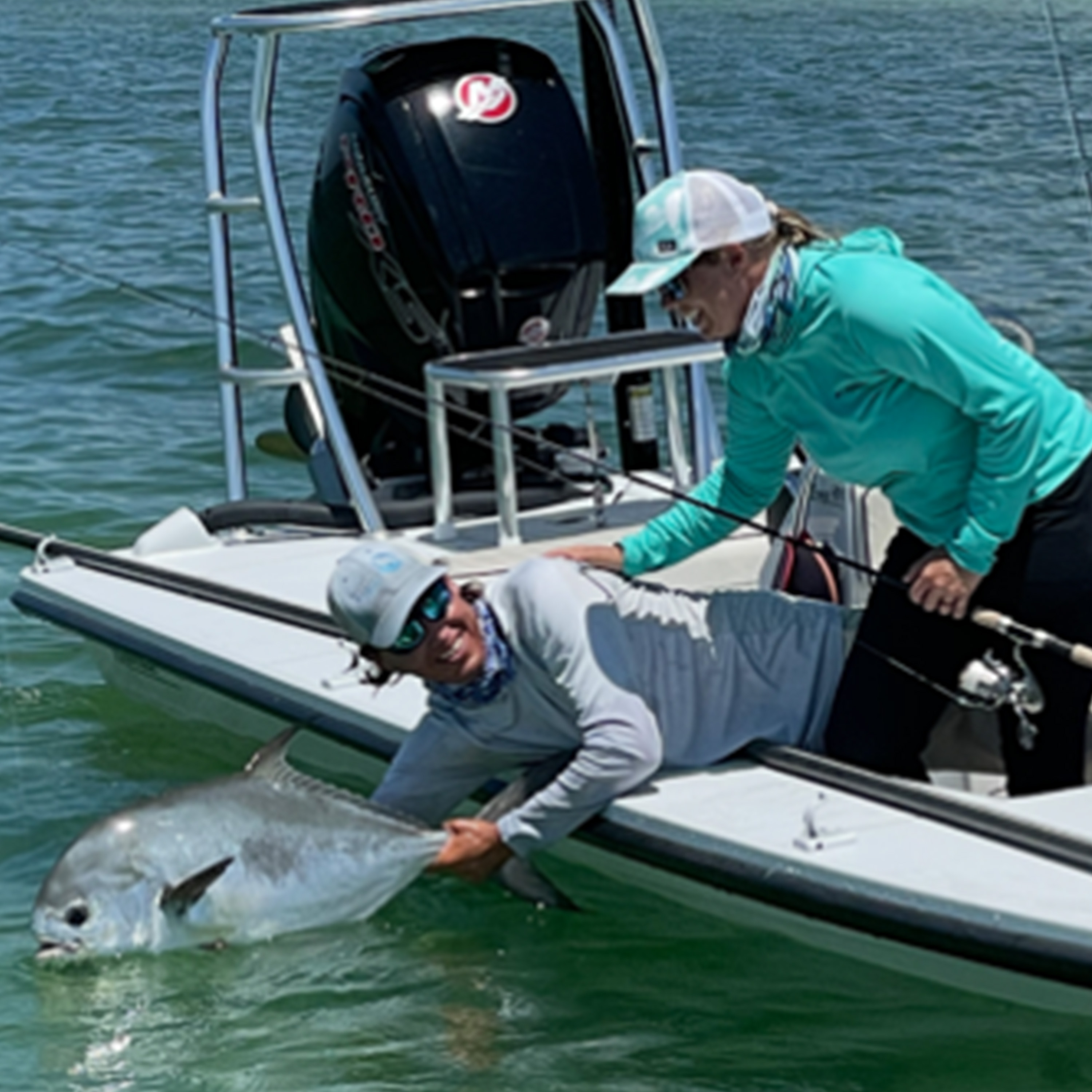 Two anglers on a fishing boat, one leaning over the side while holding a large silver-colored fish partly in green water as the other stands nearby holding a fishing rod.