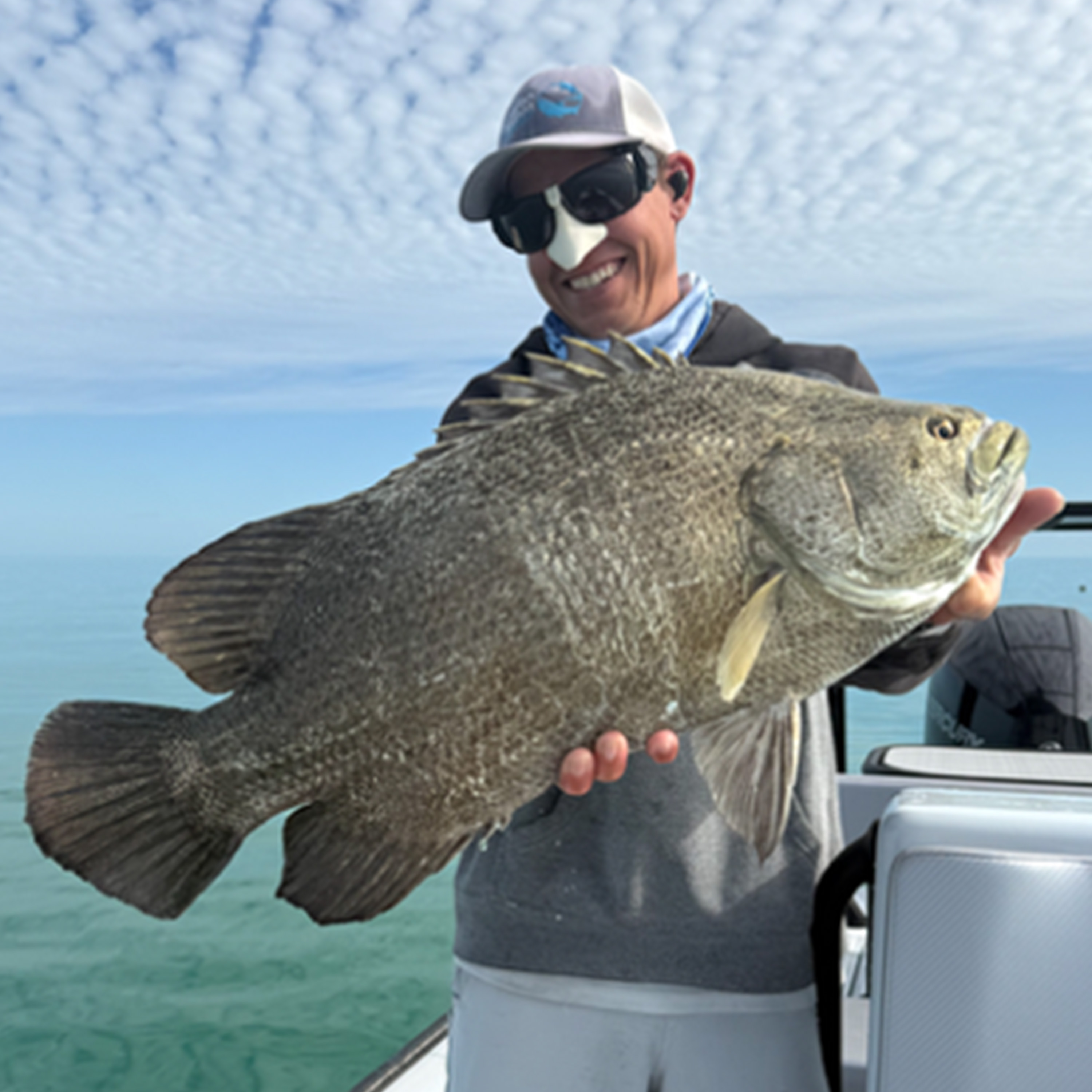 A smiling angler wearing sunglasses and a cap holds a large dark gray fish aboard a boat, with clear turquoise water and a blue, cloud-patterned sky behind him.