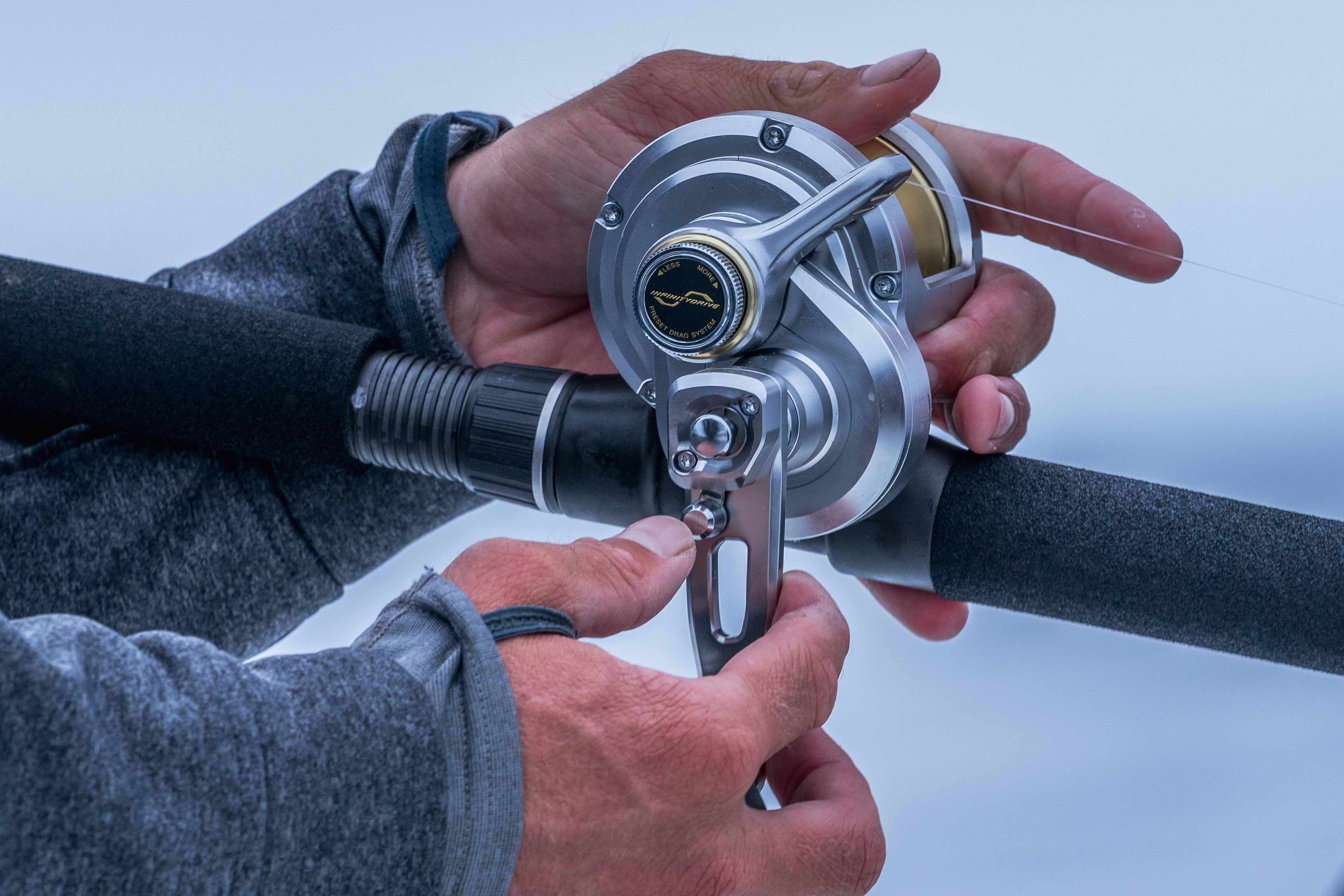 A person in a gray jacket adjusts a silver-colored conventional fishing reel mounted on a rod, with fishing line running across the reel and visible drag control details.