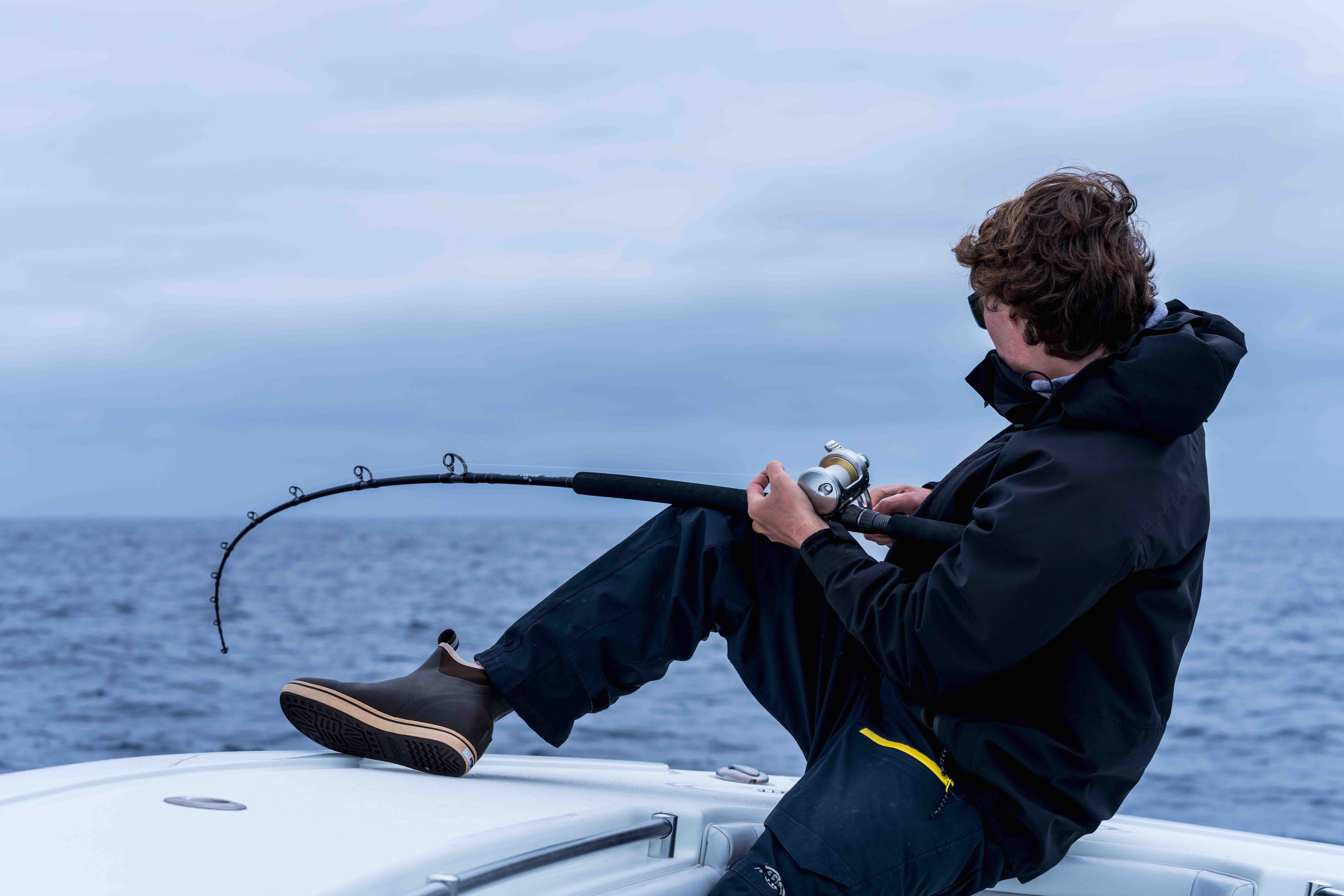 A person in black waterproof clothing sits on a boat while holding a bent fishing rod with a silver-colored conventional reel, with the ocean and cloudy sky in the background.