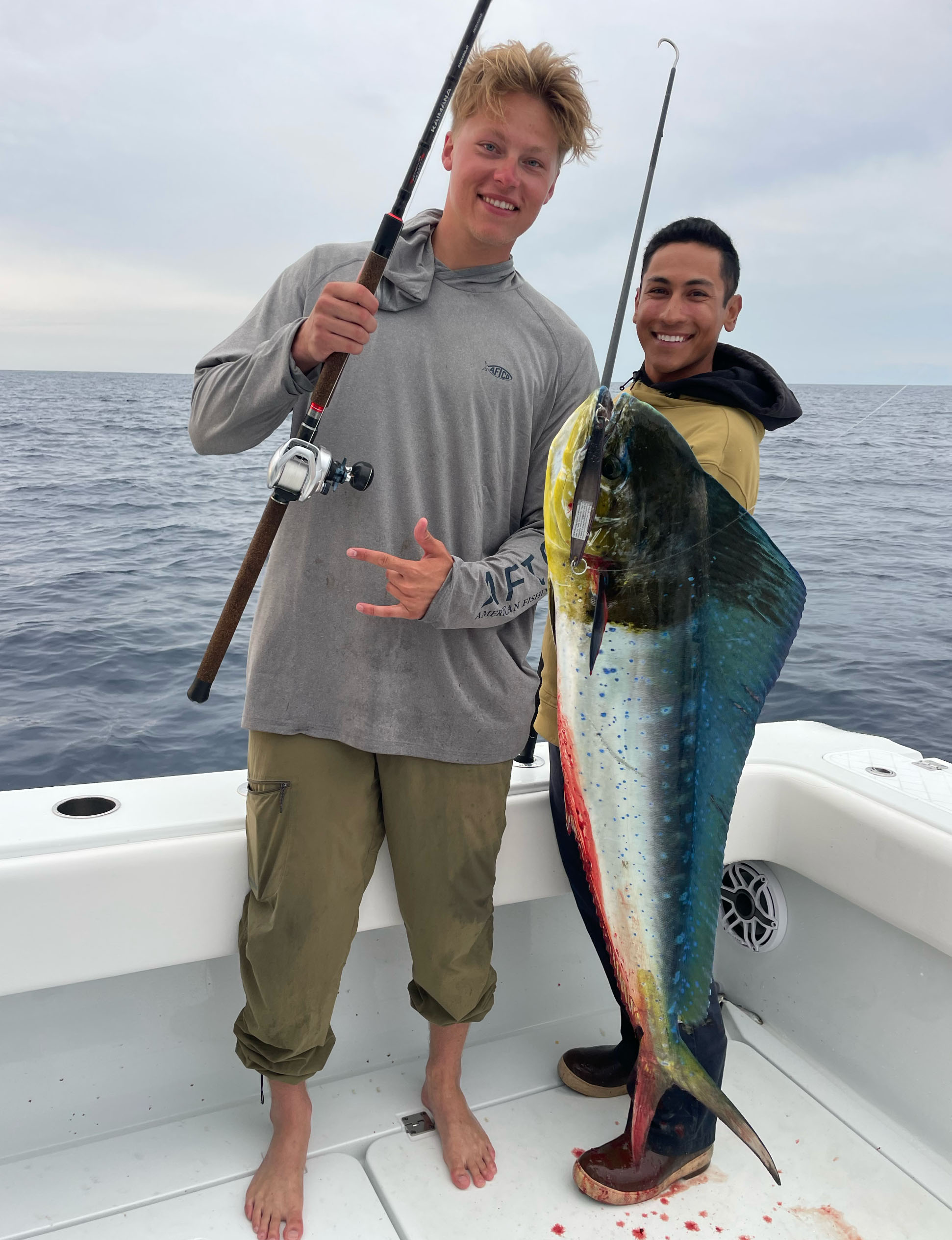 Author Ricky Fischel and another Fisherman with a Caught Dorado Fish on a boat in the ocean. The man is holding a rod and showing the Shaka hand sign on his other hand. Andy is holding a gaff. The Dorado has a lure handing from its mouth.