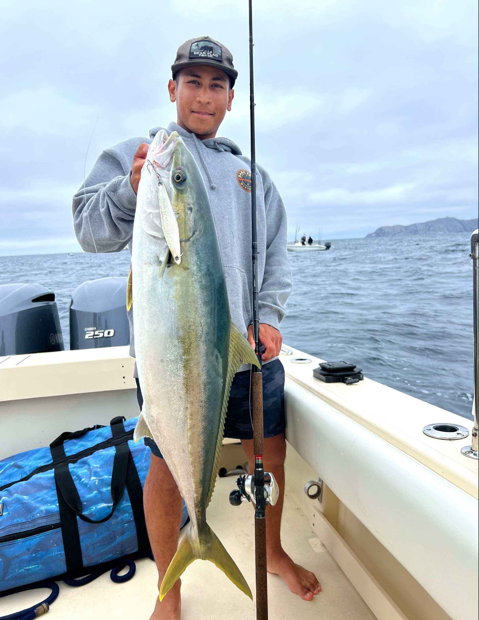 Author Ricky Fischel hands up a caught Yellowtail Fish, with his other hand on a rod leaning against his body. The fish has a lure sticking out of the mouth, hooked on a treble hook. Ricky stands on a white boat, with the ocean in the background.