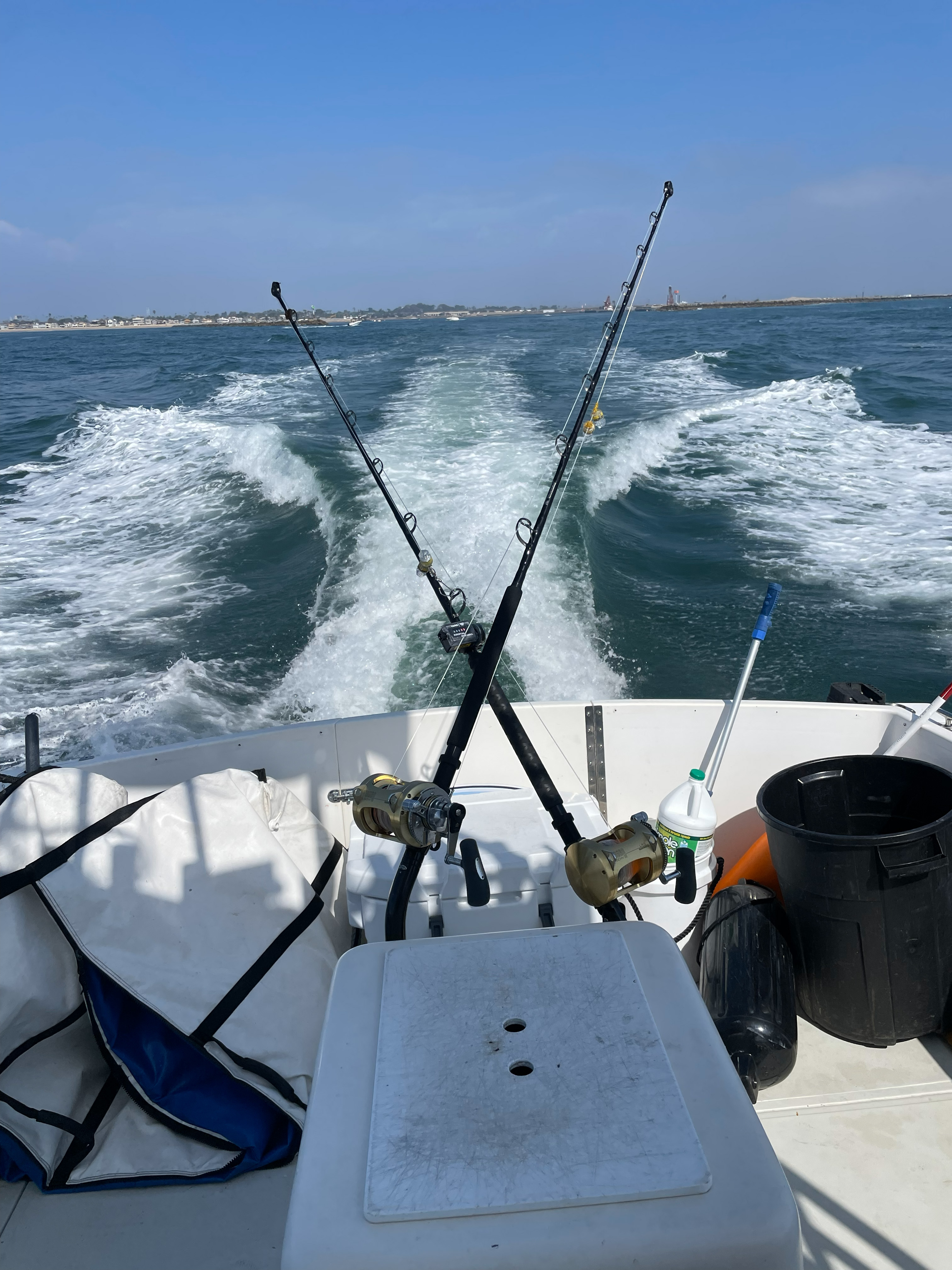 View from the stern of a boat showing two black trolling rods angled outward with gold-colored reels, lines extended into foamy wake, and deck items including a cooler and bucket.